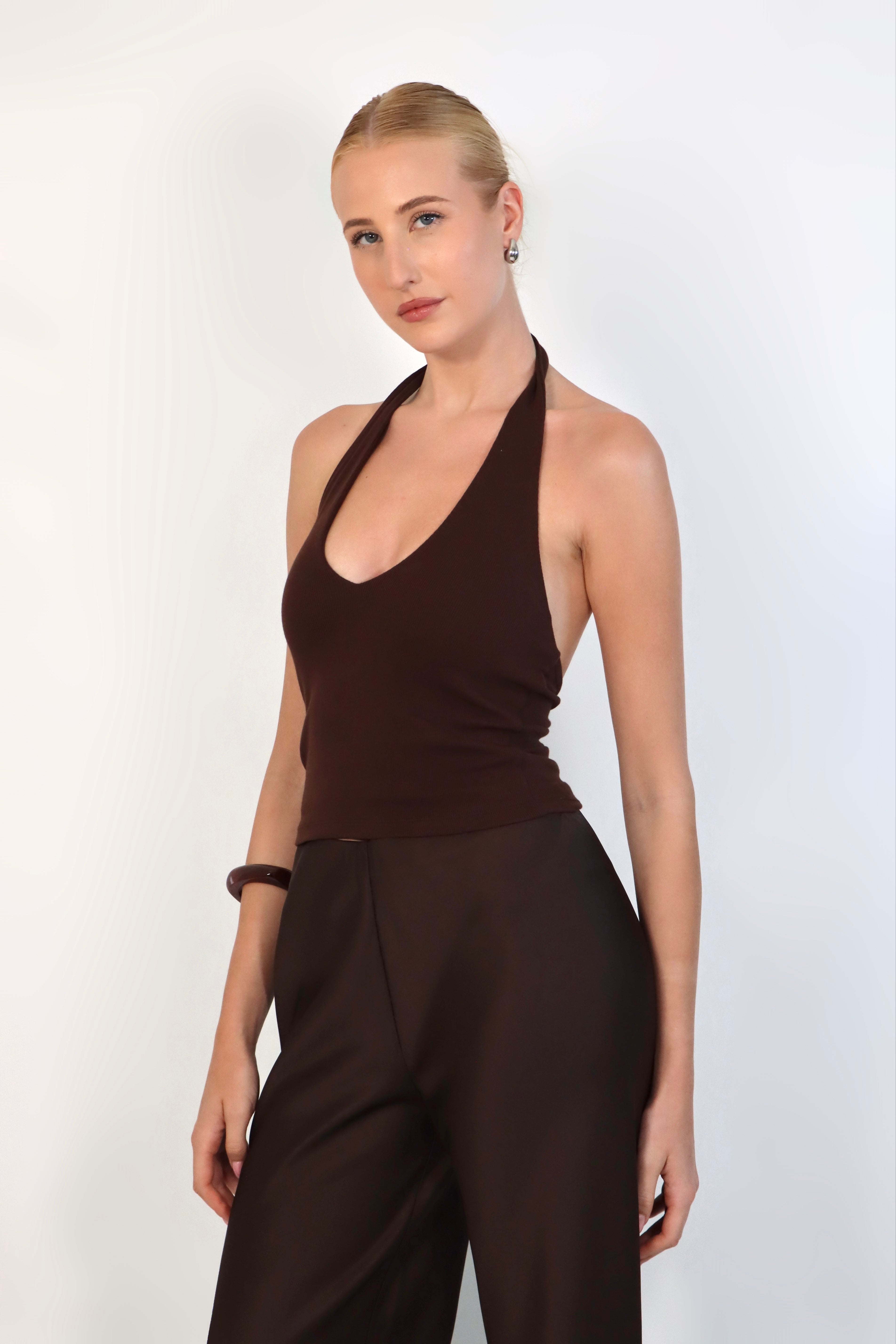A model wearing a deep Espresso halter top with a ribbed texture and a plunging V-neckline. The fitted top is styled with matching dark brown, high-waisted tailored trousers for a sleek, monochromatic look against a white studio background.