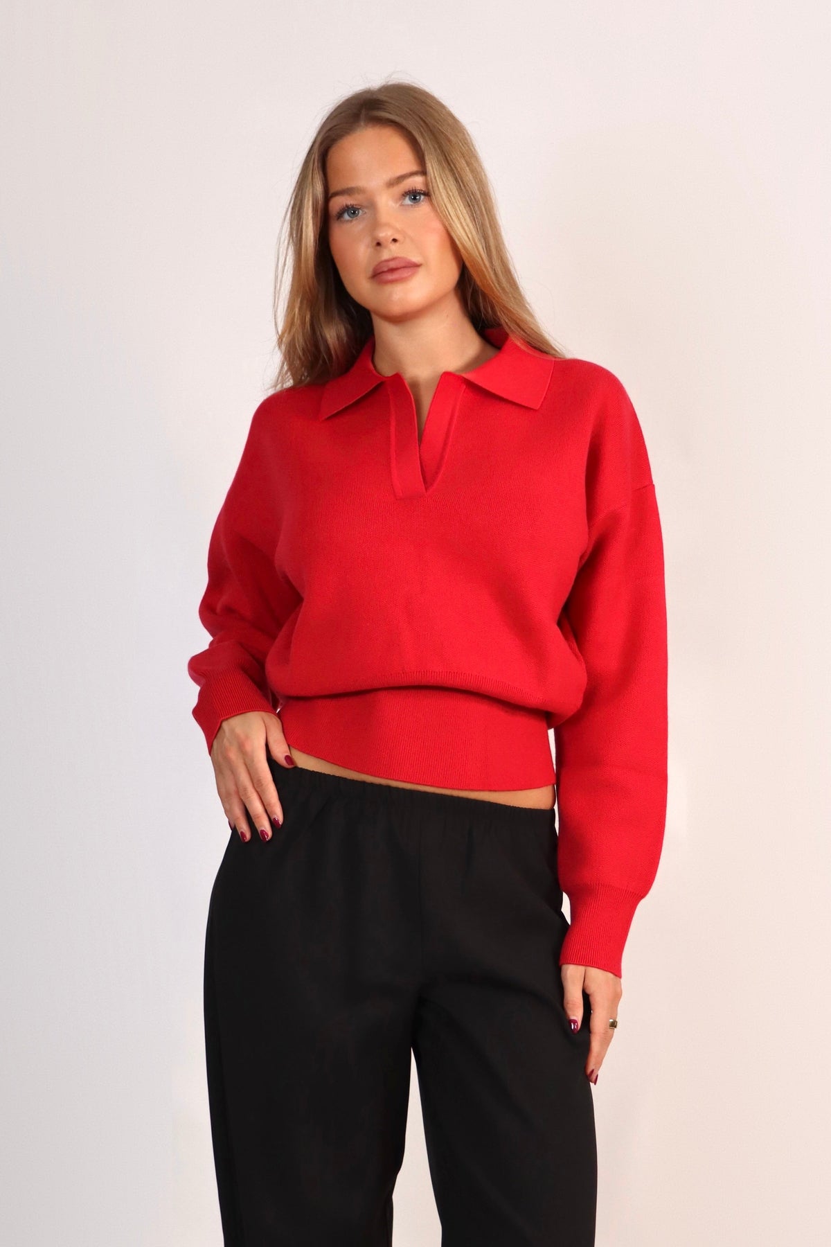 A woman modeling a bright red long-sleeve knit polo-style top with a cropped hem and black wide-leg trousers, standing against a plain light studio background.