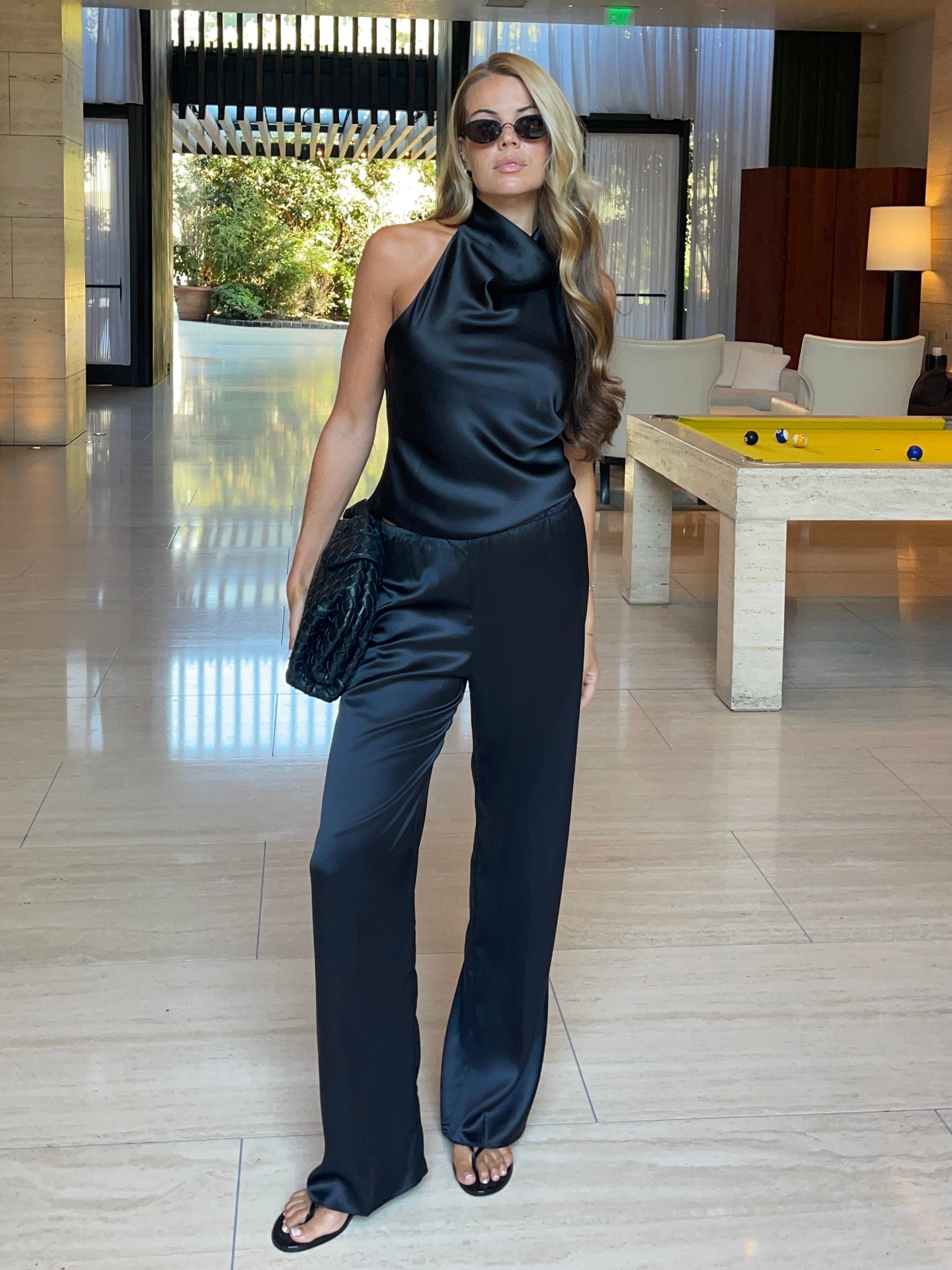 Woman wearing a black satin halter top with a draped neckline, styled with matching satin pants. The top has a smooth, glossy finish and a loose, elegant fit, creating a sophisticated and minimalist look against a light background.