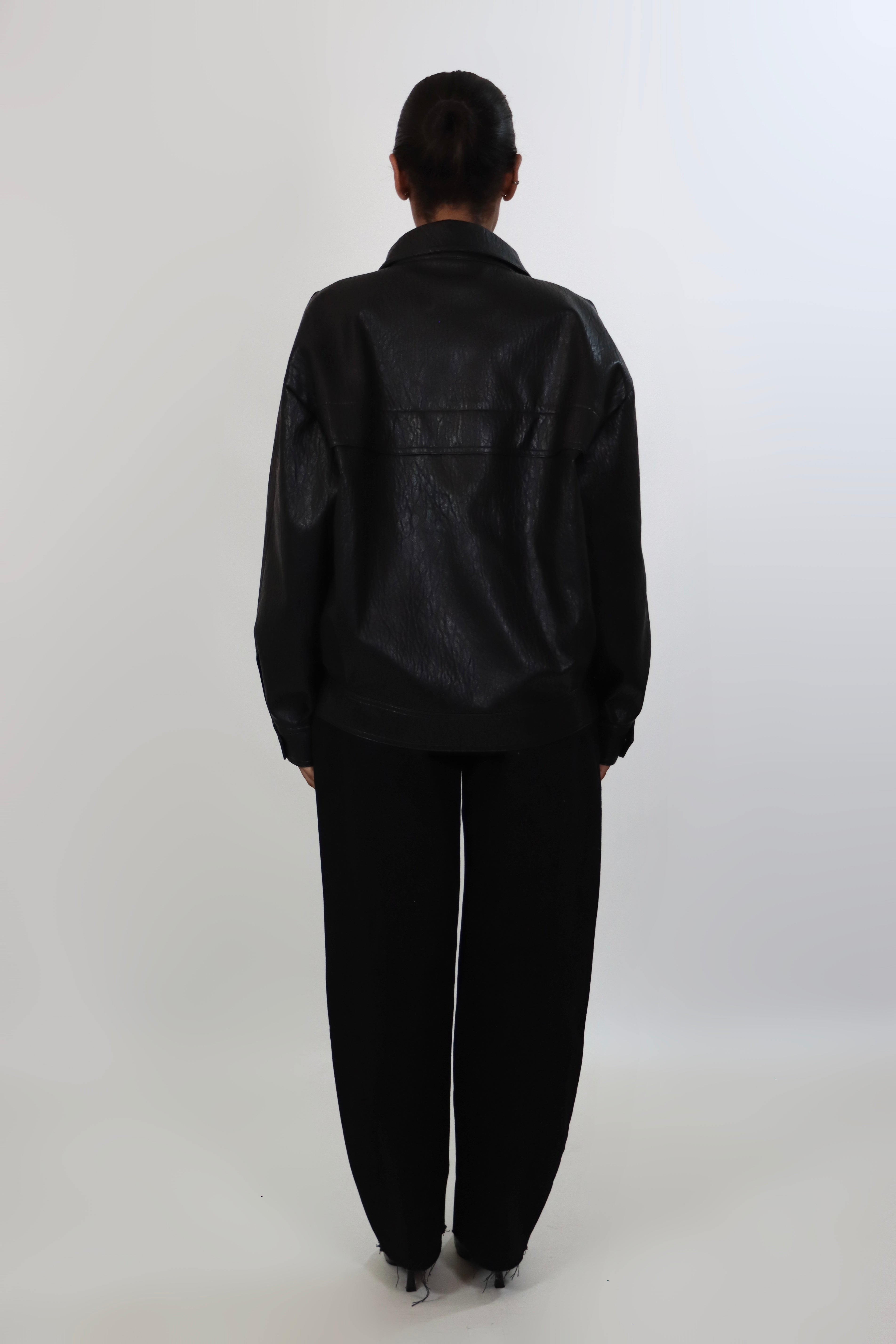 Model wearing a black textured leather oversized jacket with a structured collar and long sleeves, styled with black pants against a neutral background.