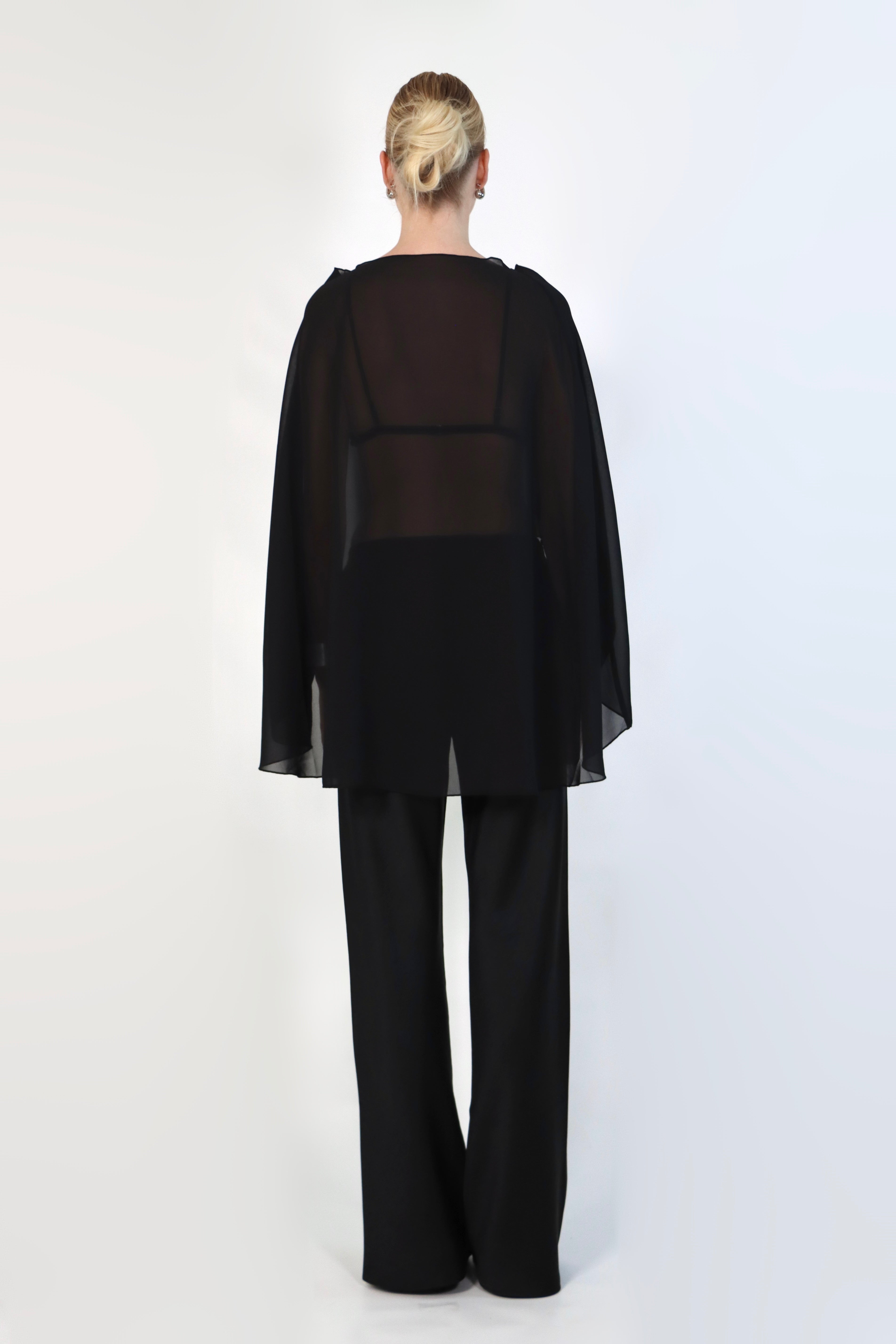 model poses against a neutral background wearing a sheer, black Chiffon Cape Top layered over a matching white bikini top. The cape features an asymmetrical, flowing drape that falls gracefully over high-waisted, cream-colored April Satin Pants and white strappy heels.