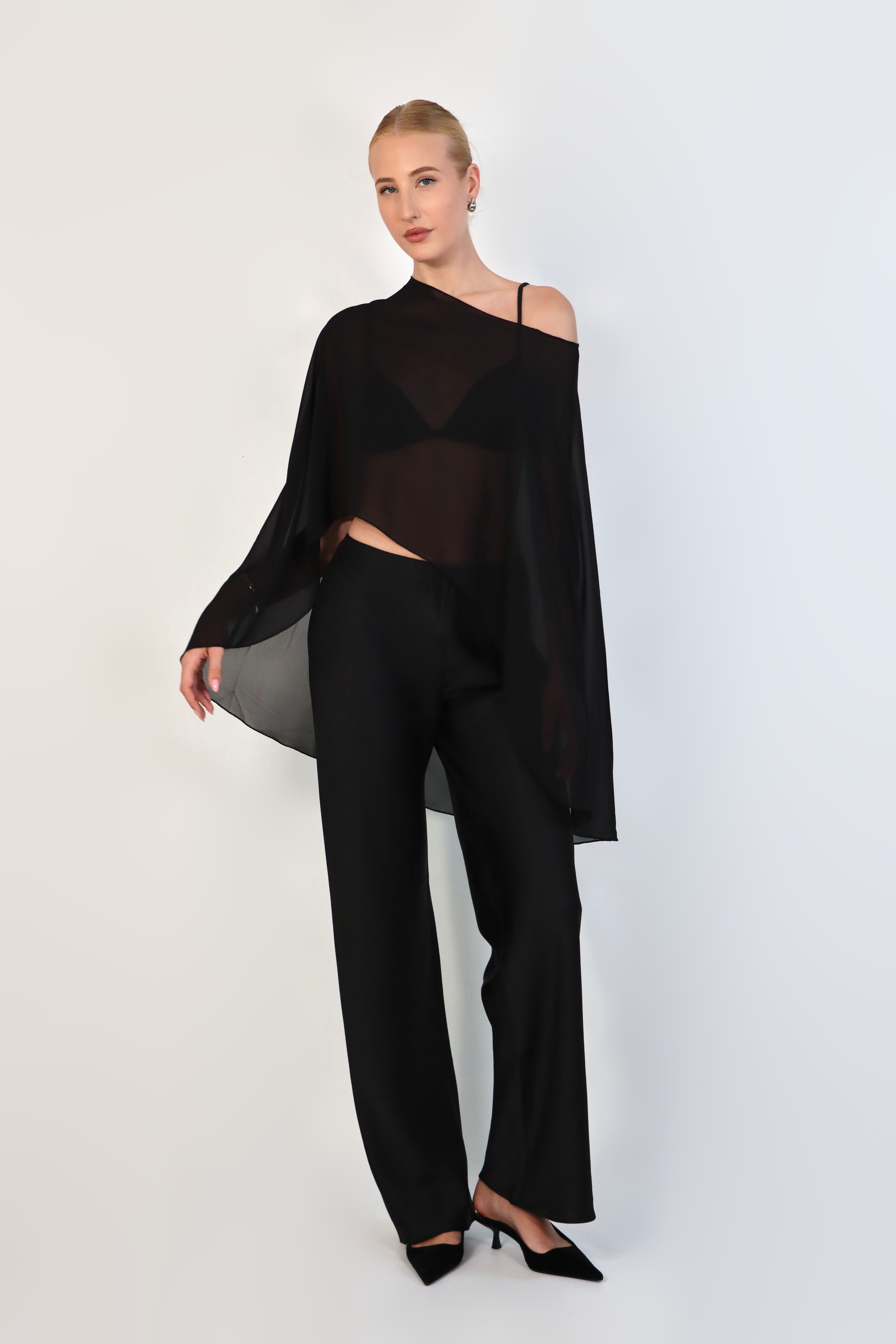 A model poses against a neutral background wearing a sheer, black Chiffon Cape Top layered over a matching white bikini top. The cape features an asymmetrical, flowing drape that falls gracefully over high-waisted, cream-colored April Satin Pants and white strappy heels.