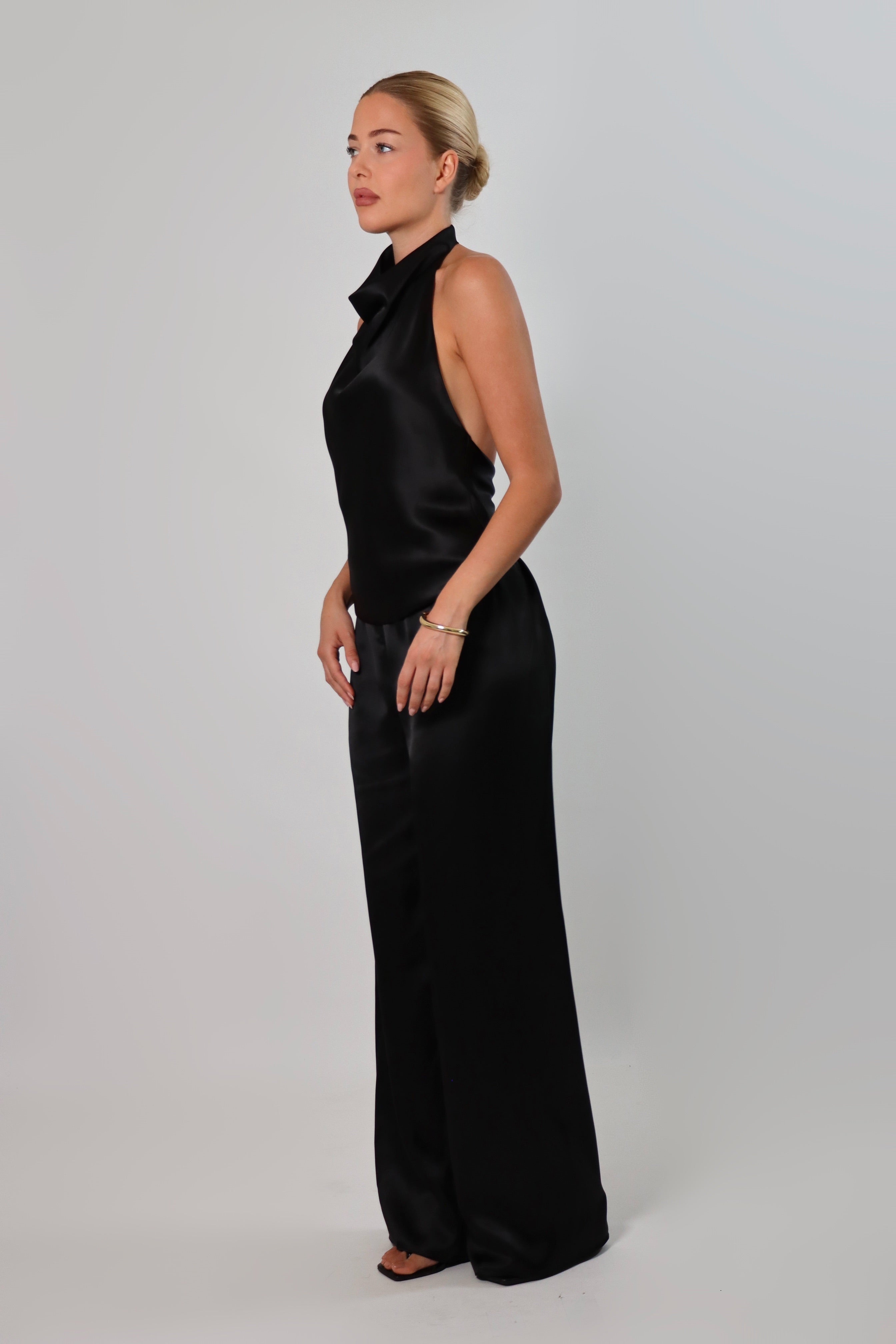 Woman wearing a black satin halter top with a draped neckline, styled with matching satin pants. The top has a smooth, glossy finish and a loose, elegant fit, creating a sophisticated and minimalist look against a light background.