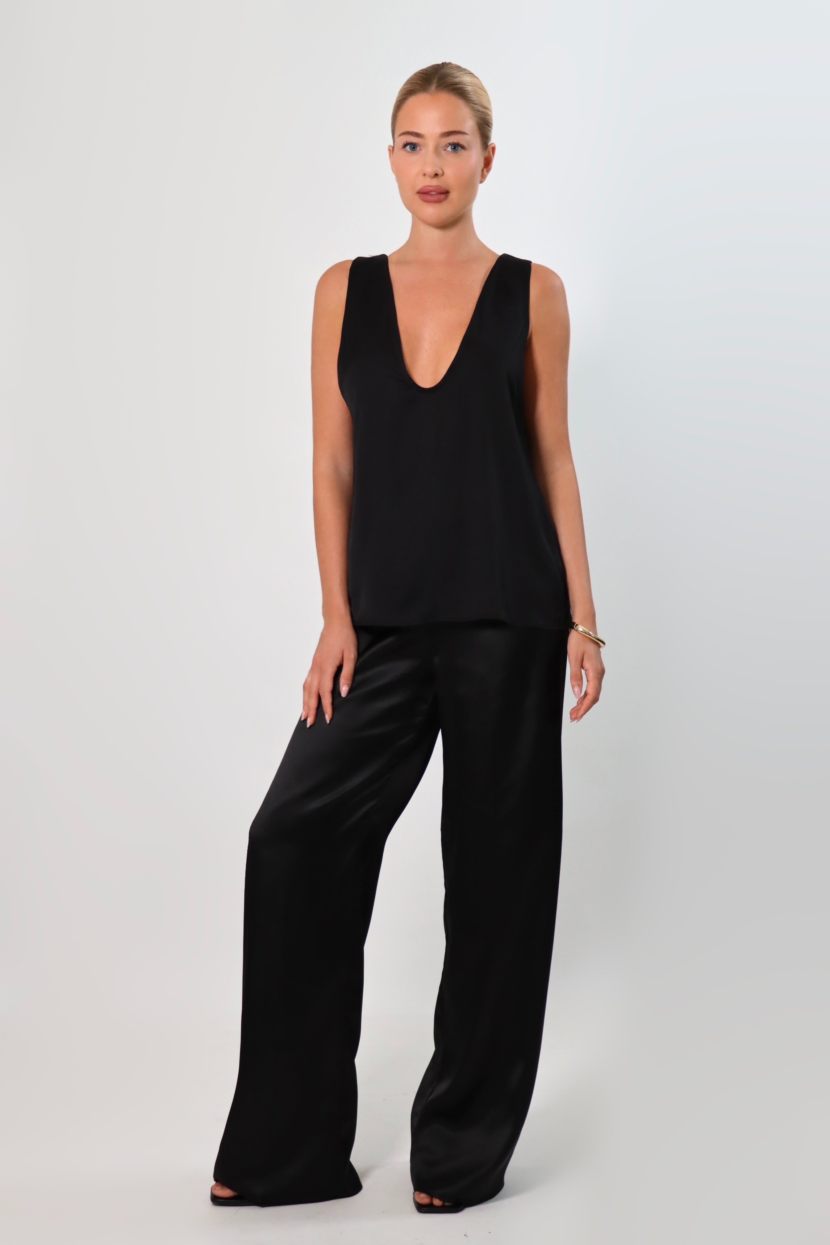 Model wearing a black satin sleeveless top with a deep plunging neckline and matching satin pants for a sleek monochrome look.