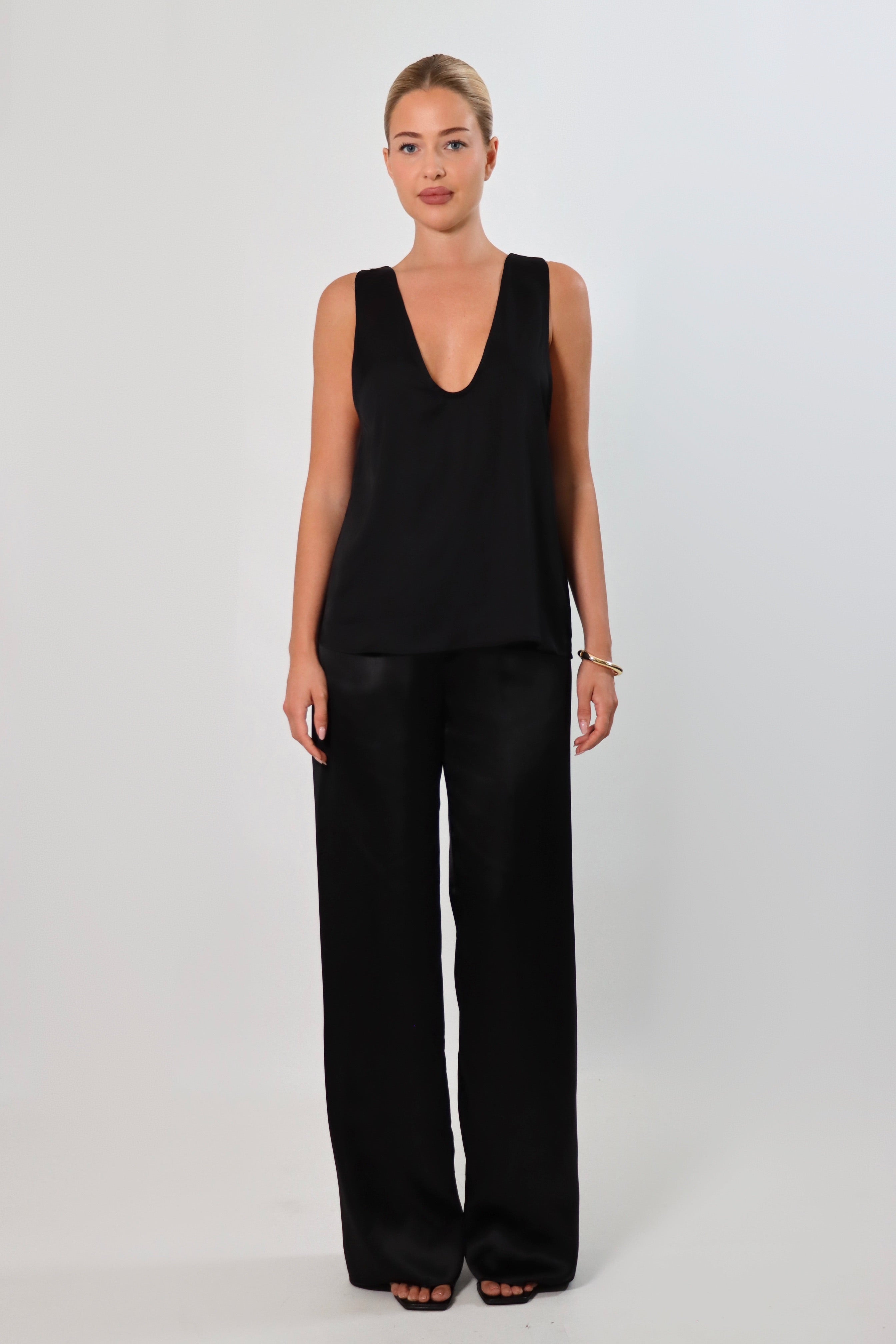Black: Model wearing a black satin sleeveless top with a deep plunging neckline and matching satin pants for a sleek monochrome look.