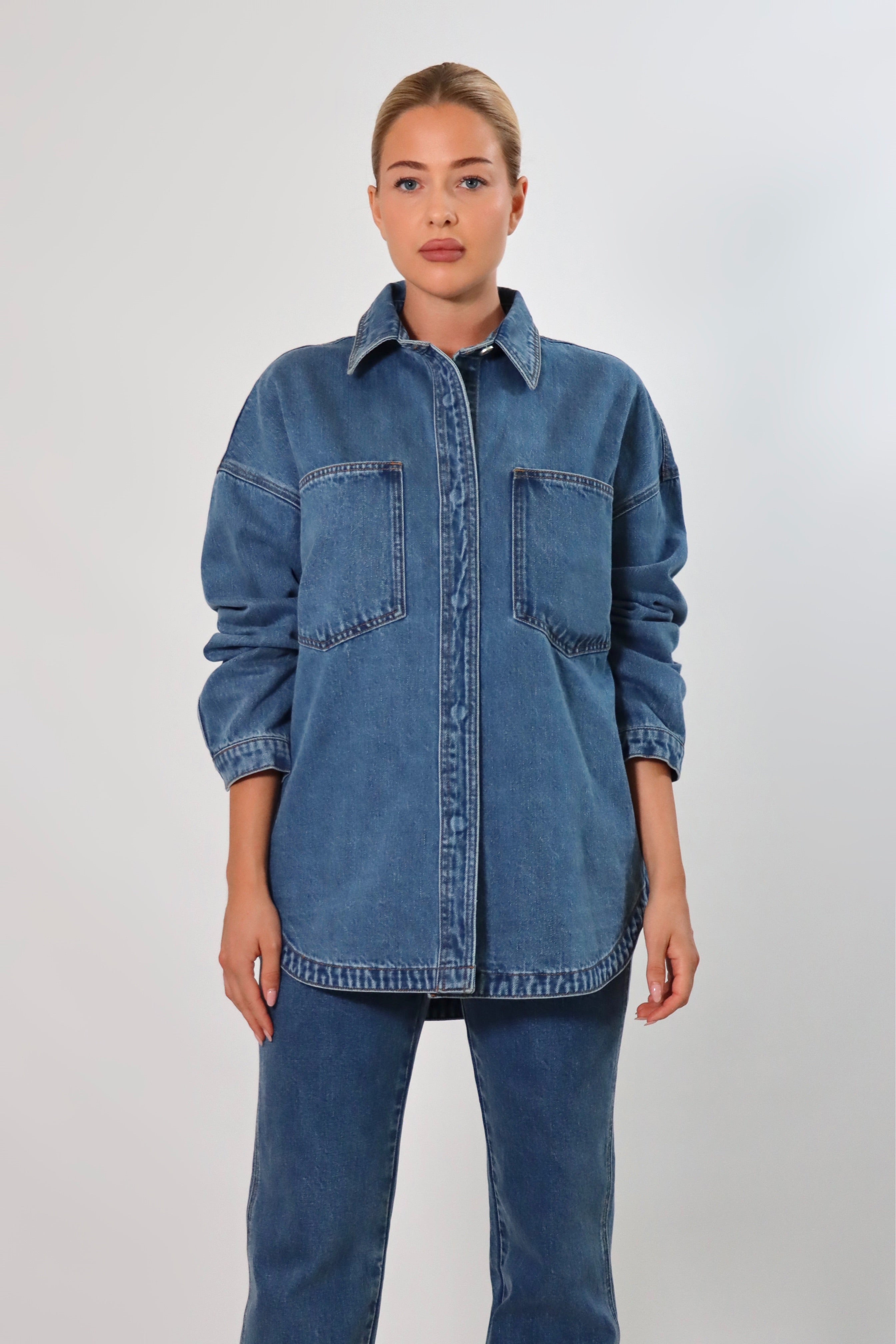 Woman wearing an oversized blue denim shirt jacket made from heavy structured denim, featuring large front pockets and long sleeves, styled with matching Stella Jeans for a full denim-on-denim look.