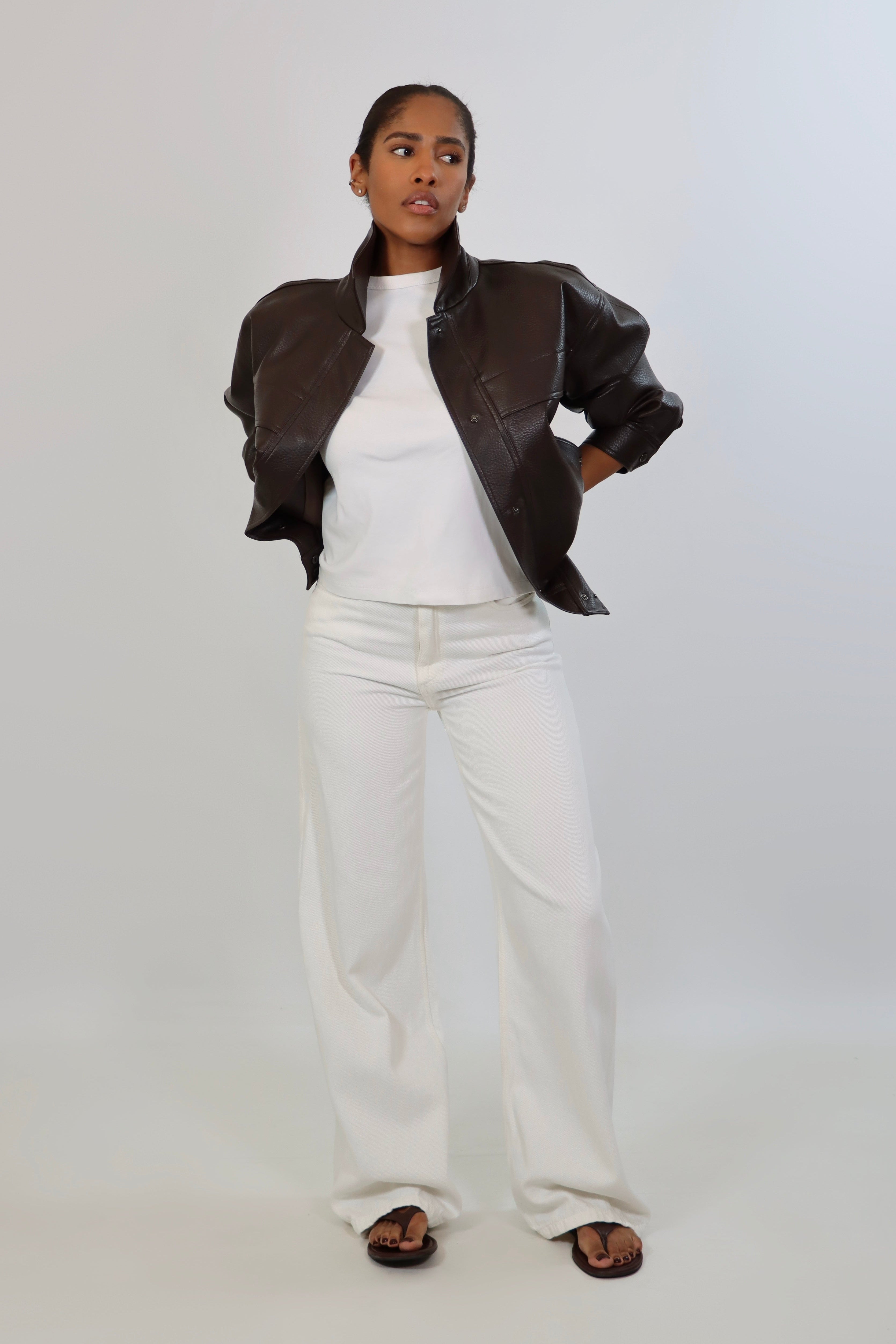 Model wearing an oversized espresso brown faux leather jacket with a boxy fit, spread collar, and front snap closure, styled with white pants against a neutral background.
