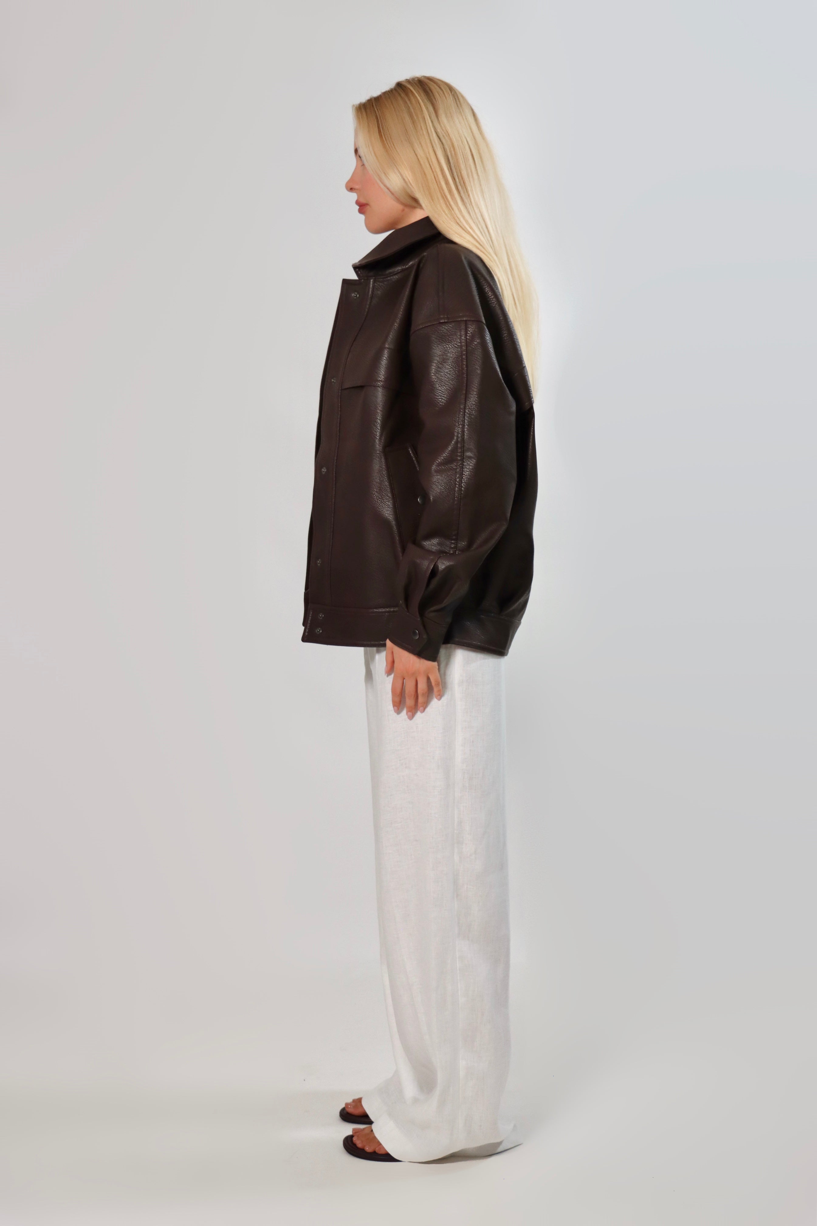 Model wearing an oversized espresso brown faux leather jacket with a boxy fit, spread collar, and front snap closure, styled with white pants against a neutral background.