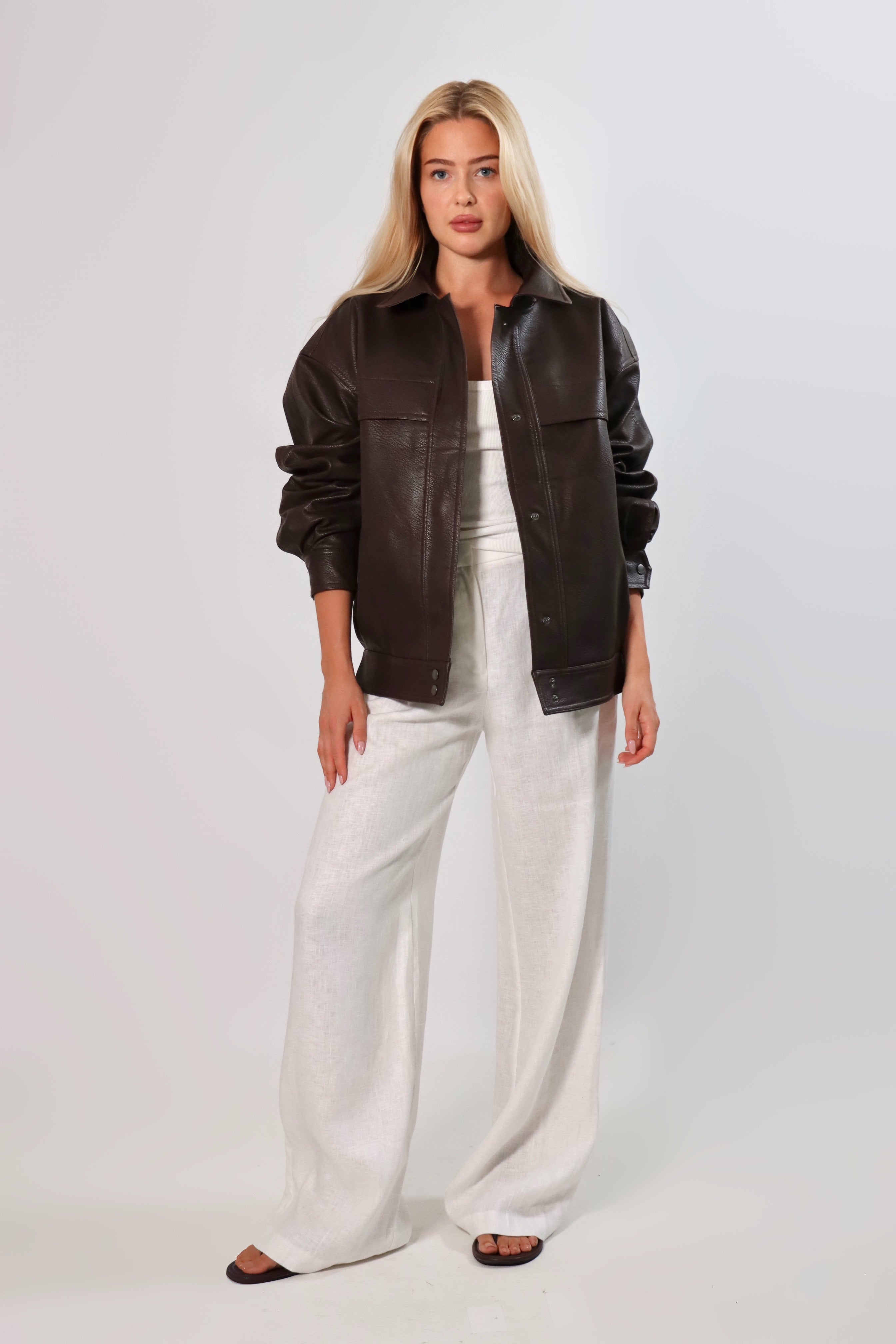 Model wearing an oversized espresso brown faux leather jacket with a boxy fit, spread collar, and front snap closure, styled with white pants against a neutral background.