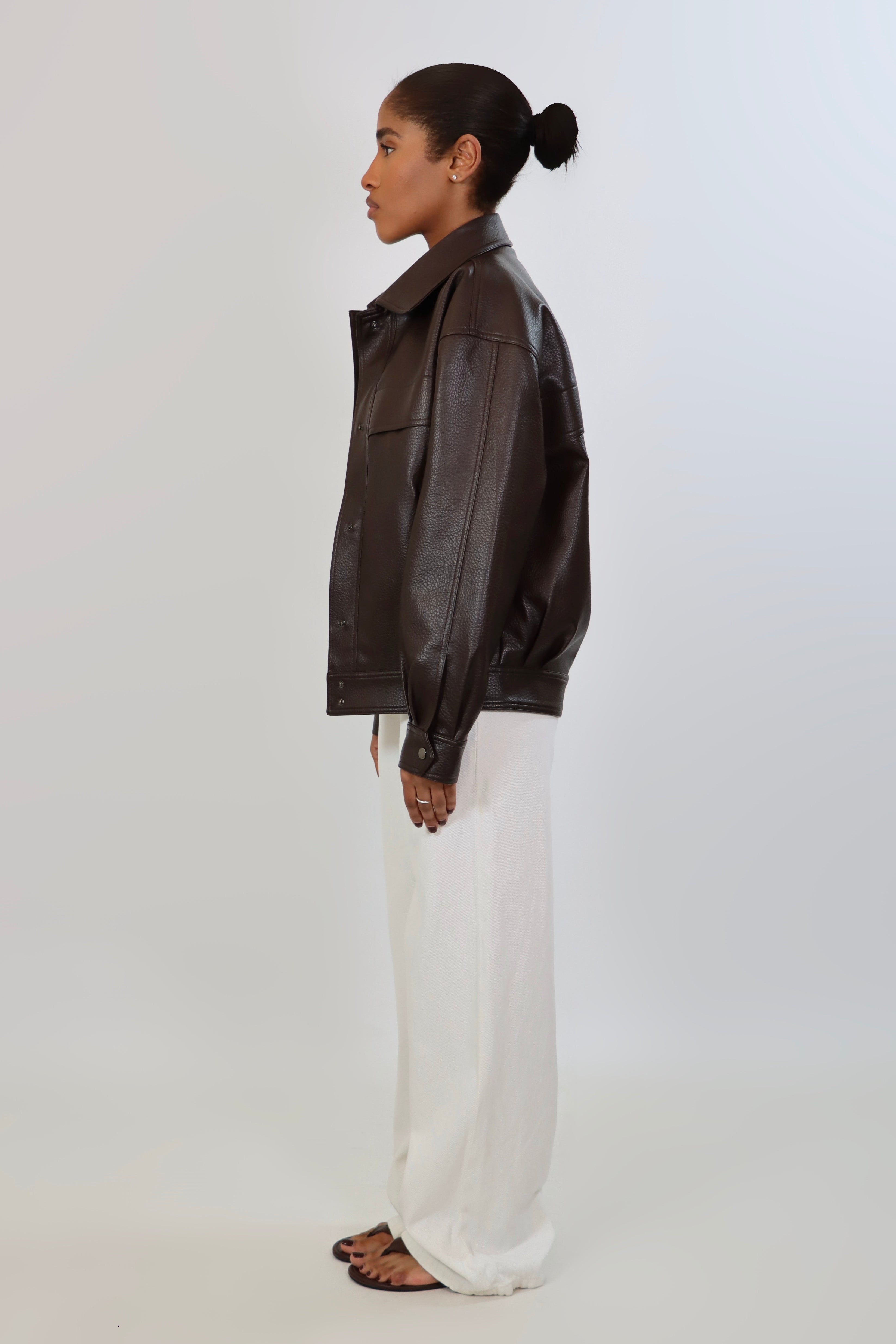 Model wearing an oversized espresso brown faux leather jacket with a boxy fit, spread collar, and front snap closure, styled with white pants against a neutral background.