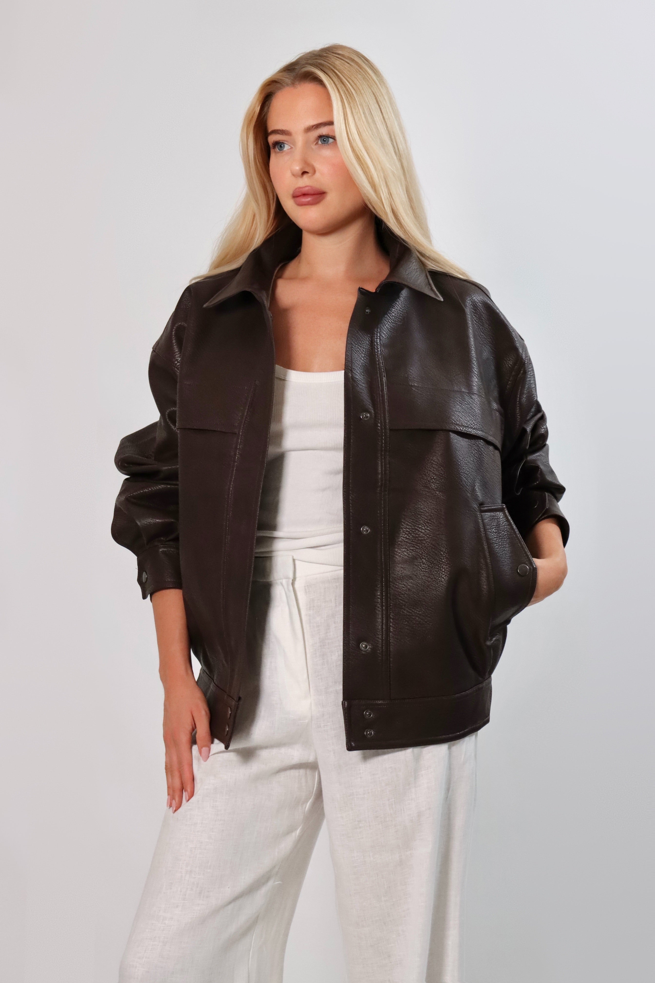 Model wearing an oversized espresso brown faux leather jacket with a boxy fit, spread collar, and front snap closure, styled with white pants against a neutral background.