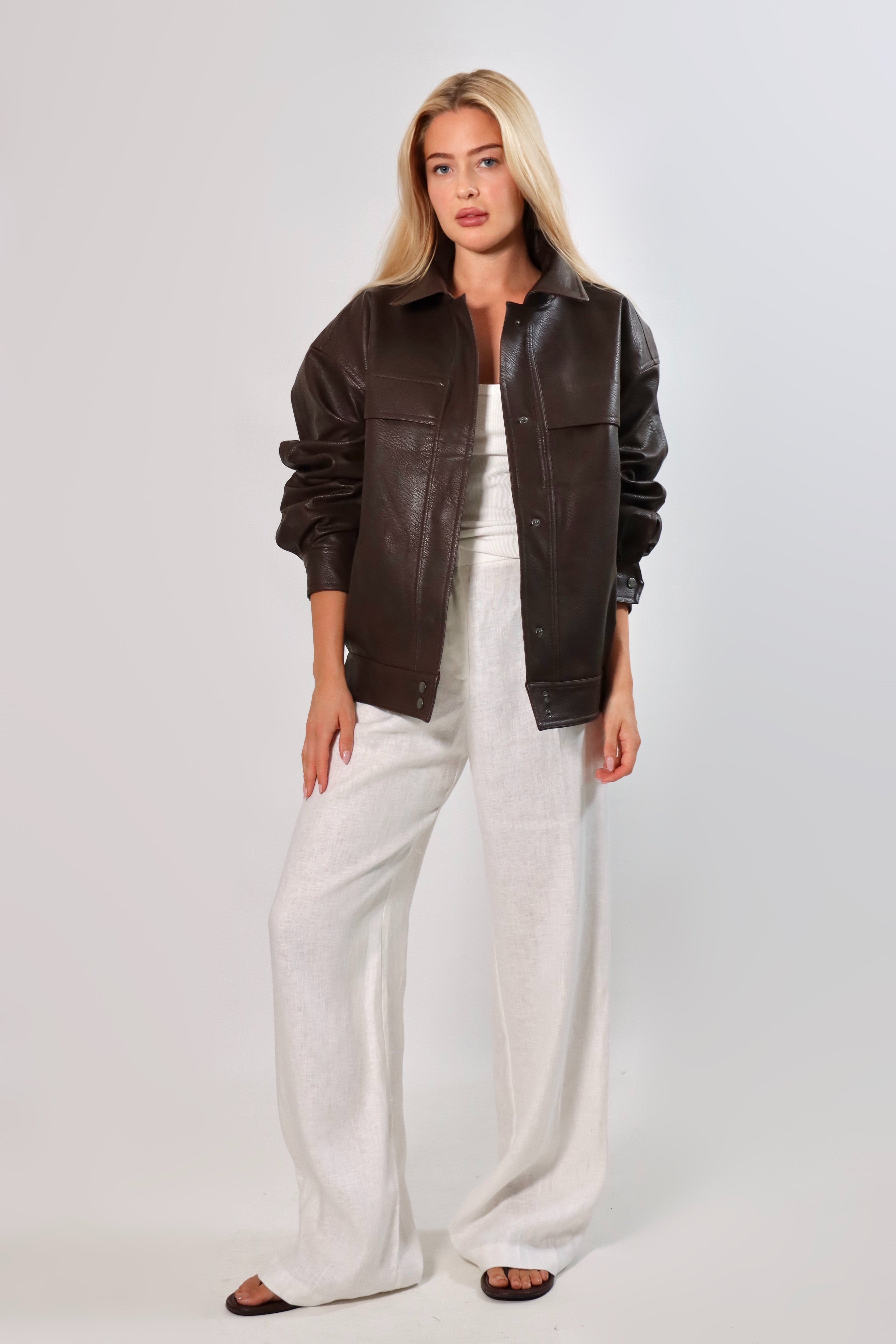 Model wearing an oversized espresso brown faux leather jacket with a boxy fit, spread collar, and front snap closure, styled with white pants against a neutral background.
