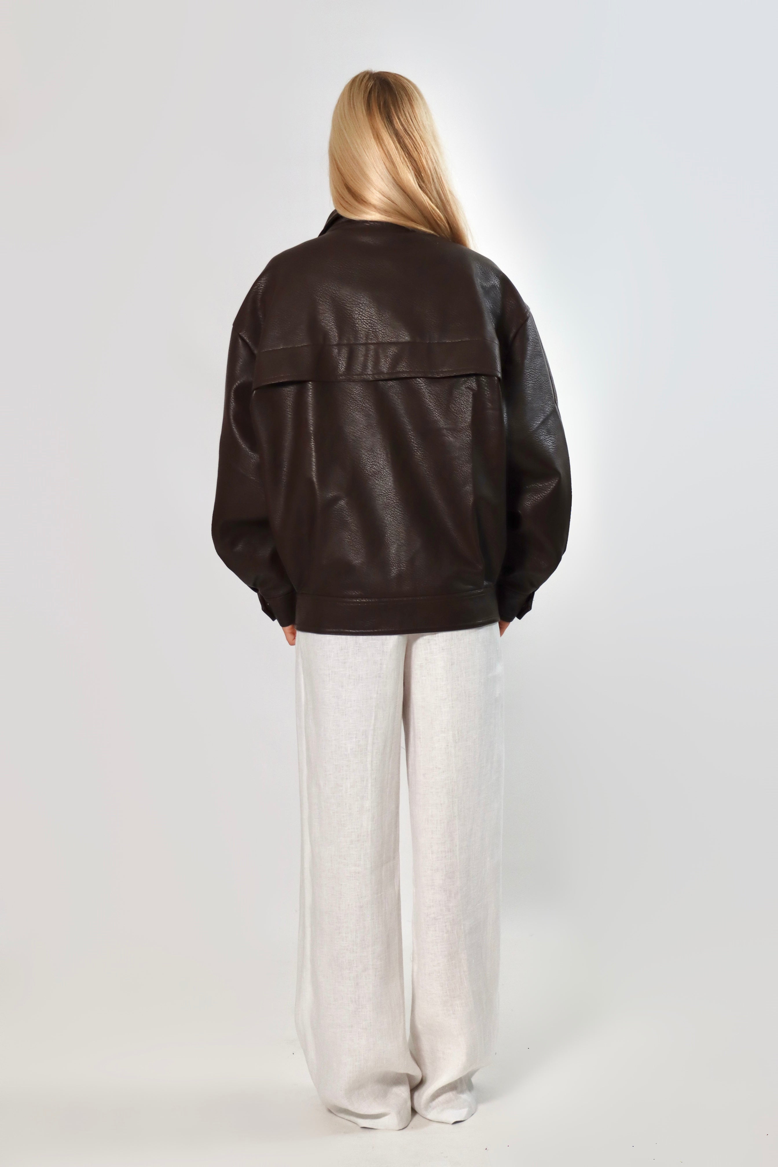 Model wearing an oversized espresso brown faux leather jacket with a boxy fit, spread collar, and front snap closure, styled with white pants against a neutral background.