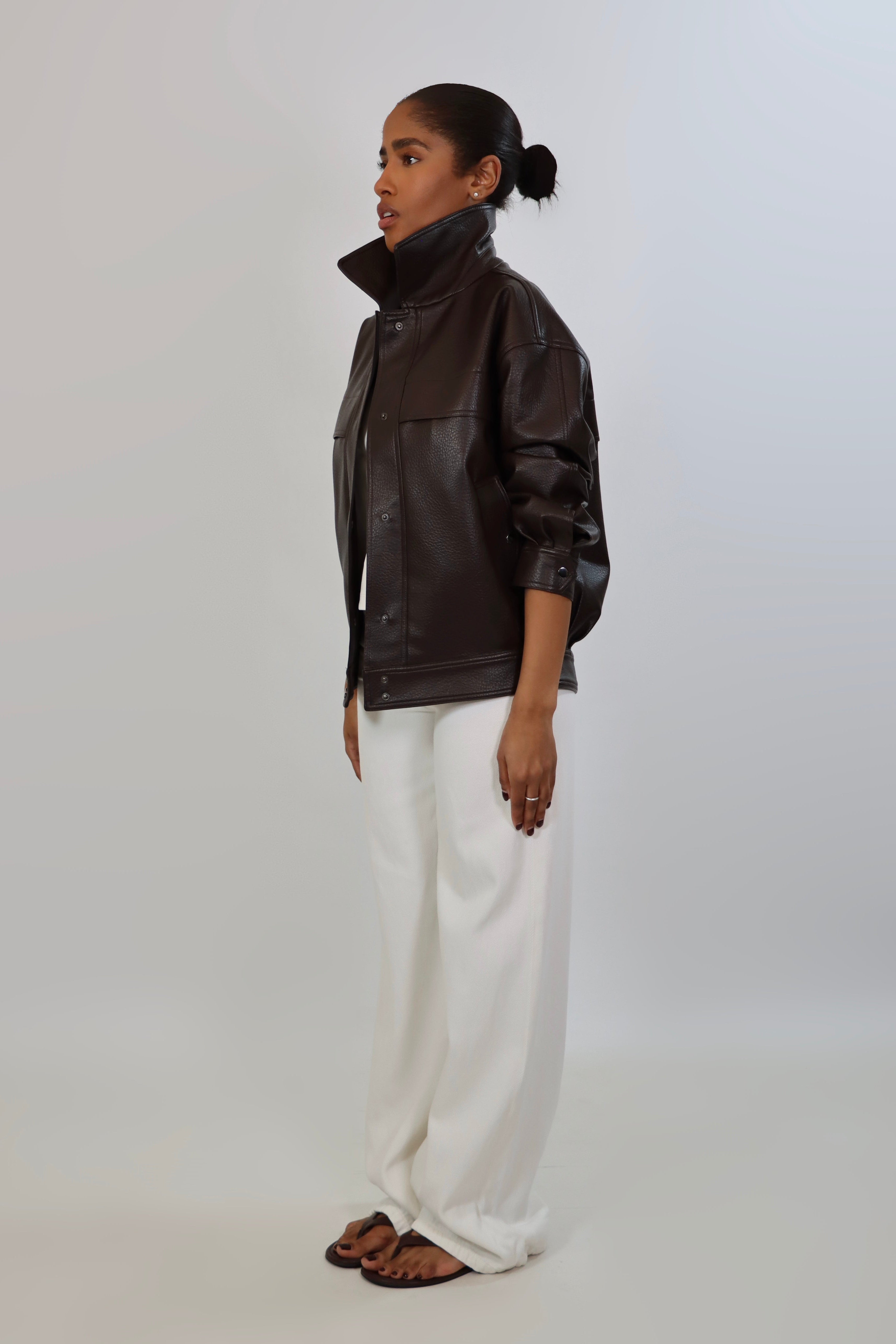 Model wearing an oversized espresso brown faux leather jacket with a boxy fit, spread collar, and front snap closure, styled with white pants against a neutral background.