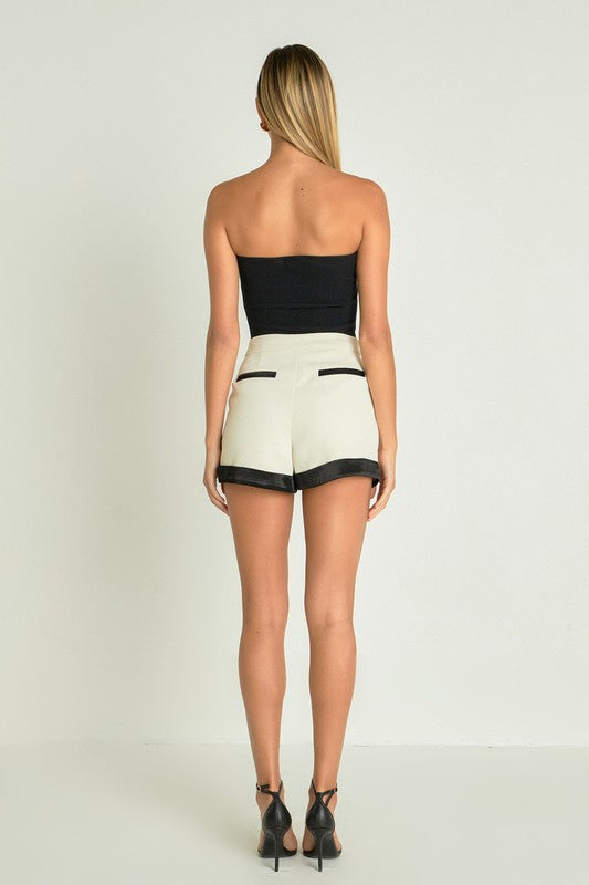 Back view: Woman in black top and cream high-waisted shorts with black satin trim and pockets, wearing black heels.