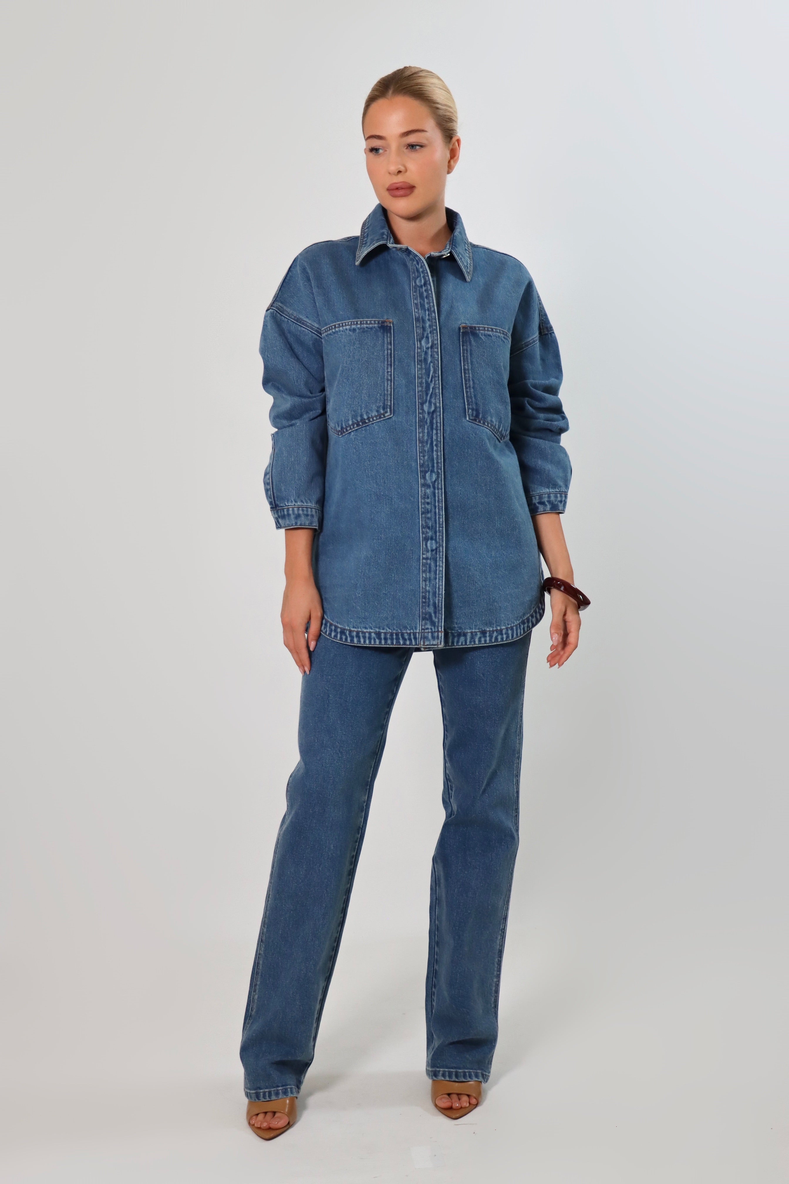 Woman wearing an oversized blue denim shirt jacket made from heavy structured denim, featuring large front pockets and long sleeves, styled with matching Stella Jeans for a full denim-on-denim look.