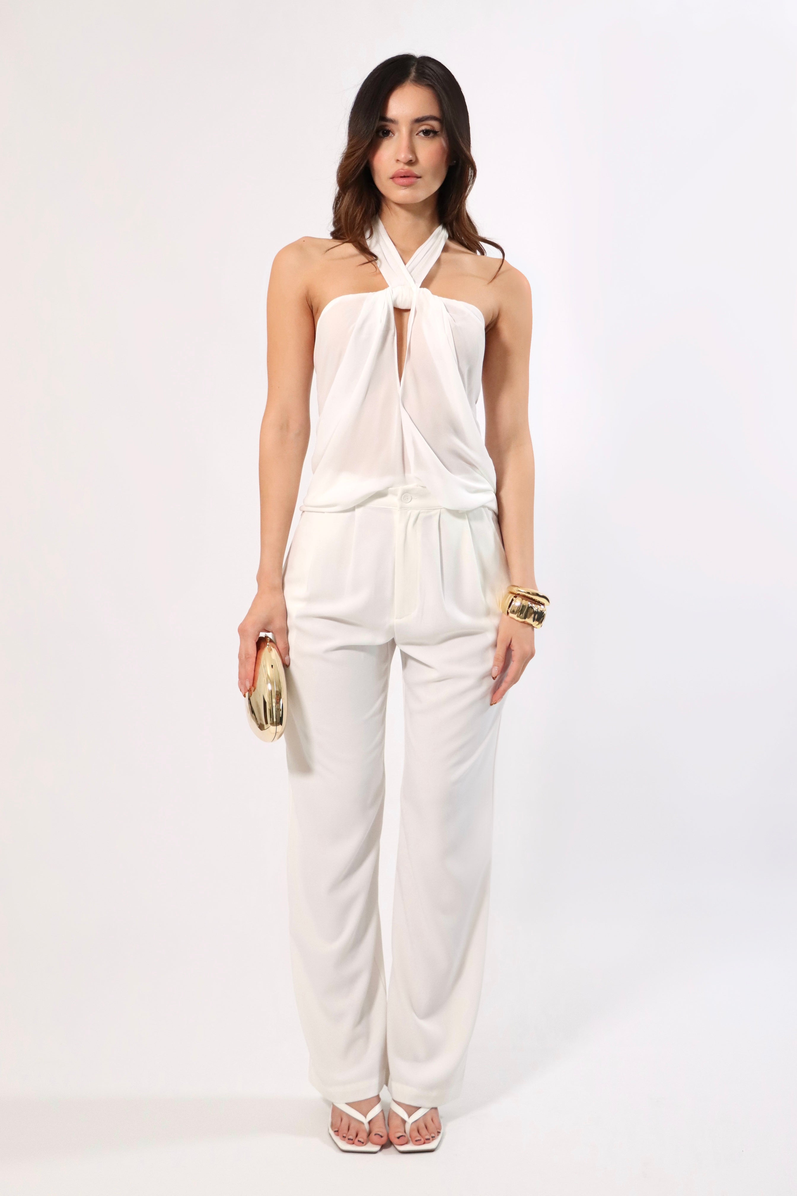 Woman in white halter top and wide-leg pants, holding gold clutch and wearing gold cuff bracelets.