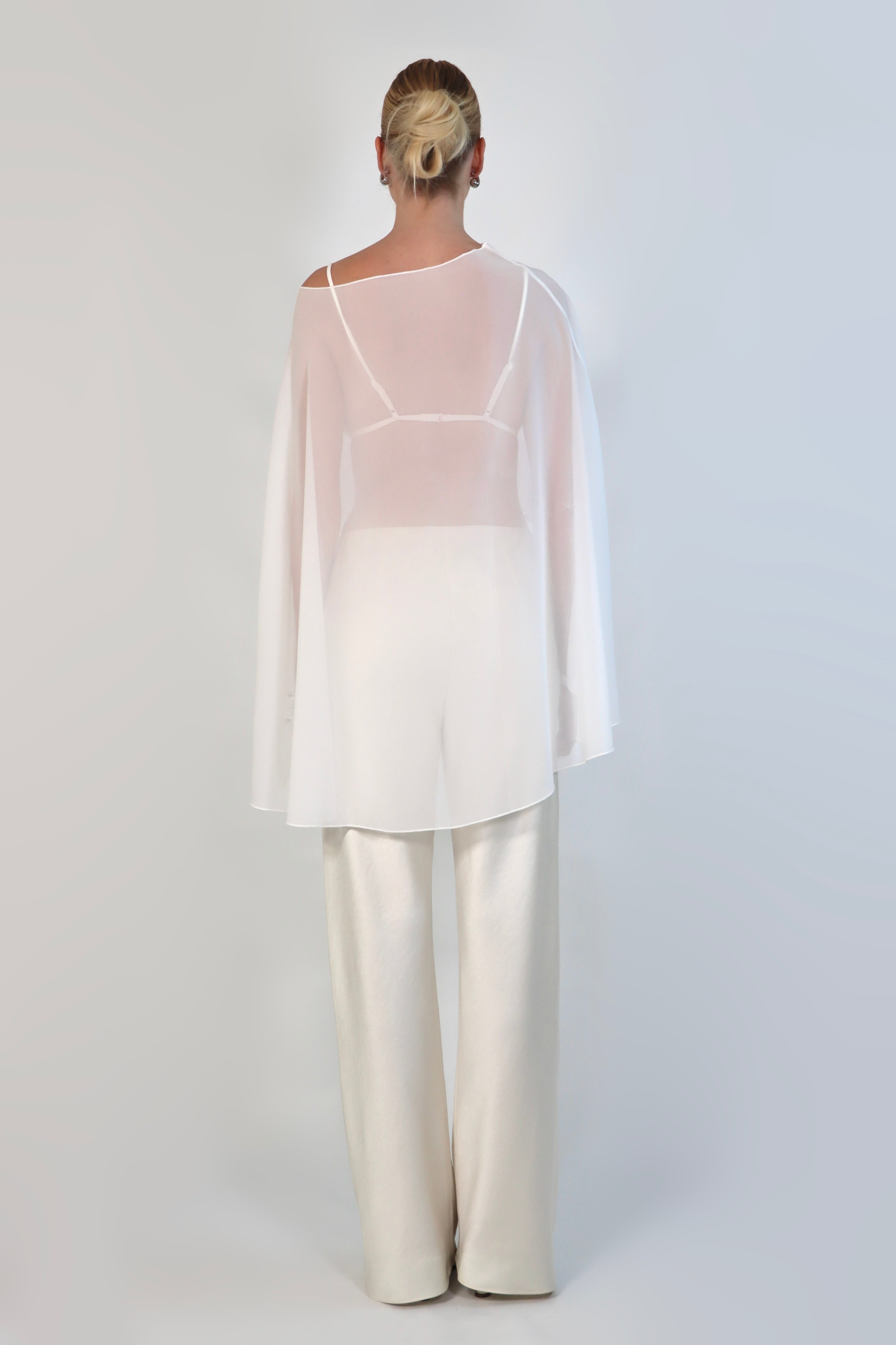 A model poses against a neutral background wearing a sheer, white Chiffon Cape Top layered over a matching white bikini top. The cape features an asymmetrical, flowing drape that falls gracefully over high-waisted, cream-colored April Satin Pants and white strappy heels.