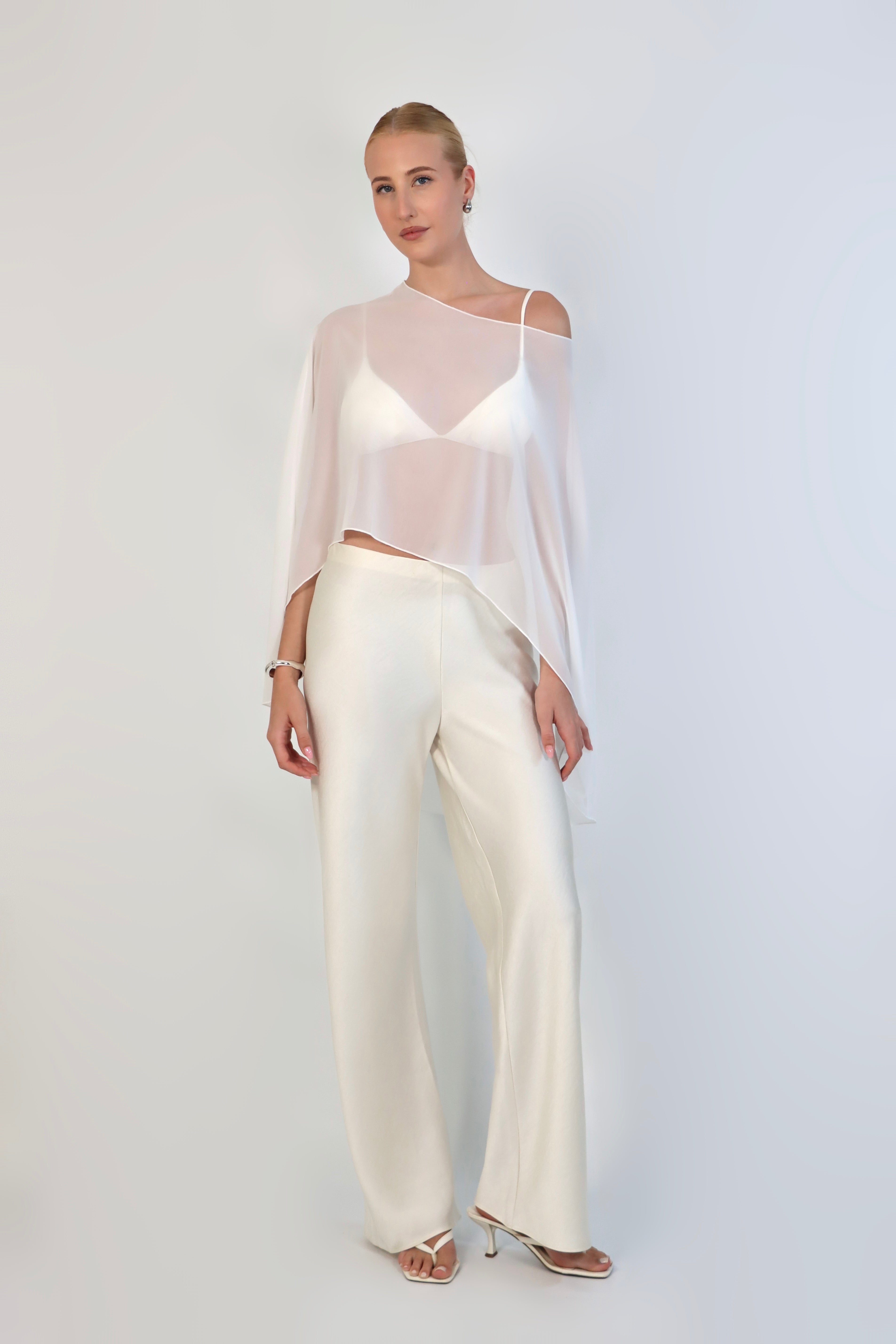 A model poses against a neutral background wearing a sheer, white Chiffon Cape Top layered over a matching white bikini top. The cape features an asymmetrical, flowing drape that falls gracefully over high-waisted, cream-colored April Satin Pants and white strappy heels.