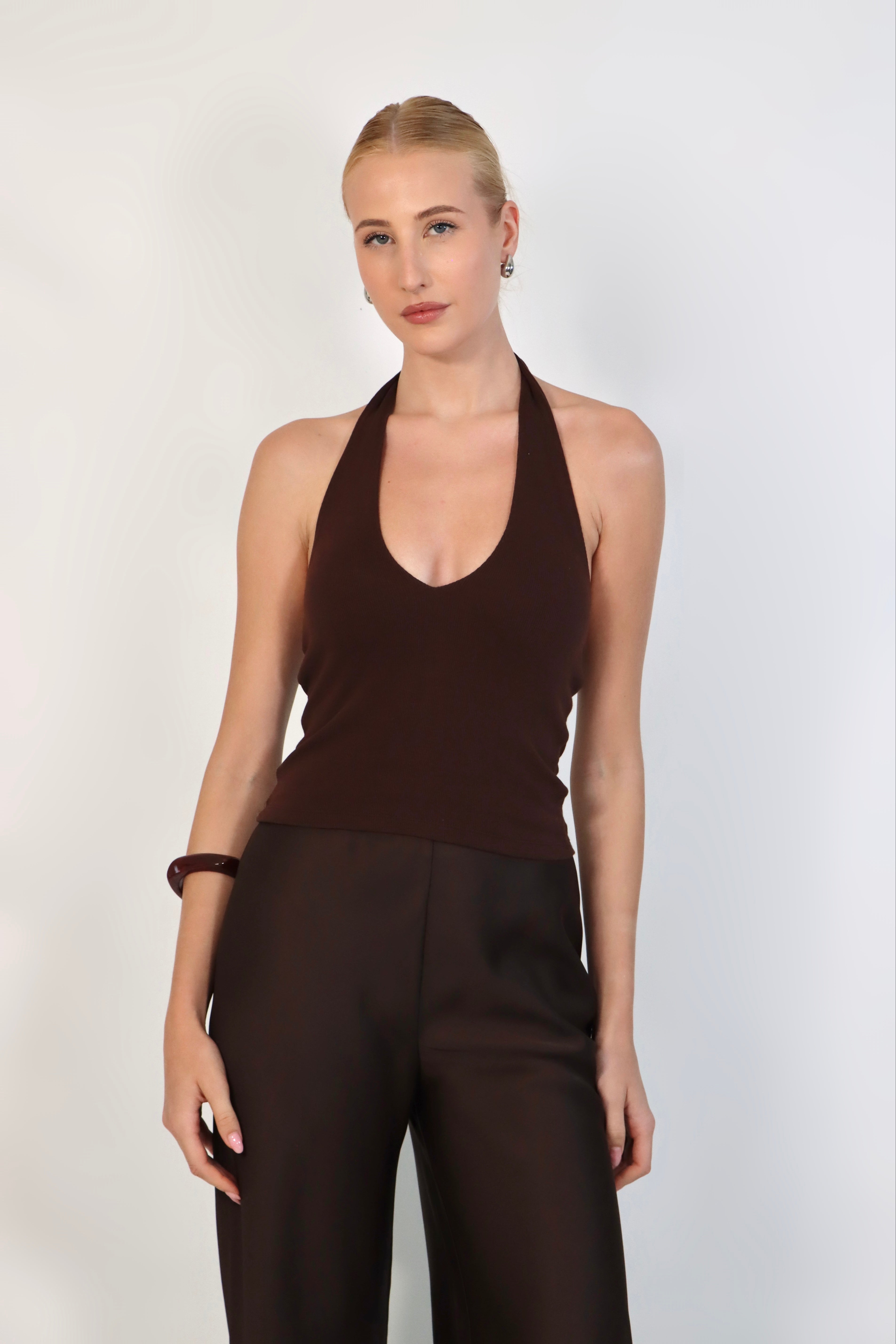 A model wearing a deep Espresso halter top with a ribbed texture and a plunging V-neckline. The fitted top is styled with matching dark brown, high-waisted tailored trousers for a sleek, monochromatic look against a white studio background.
