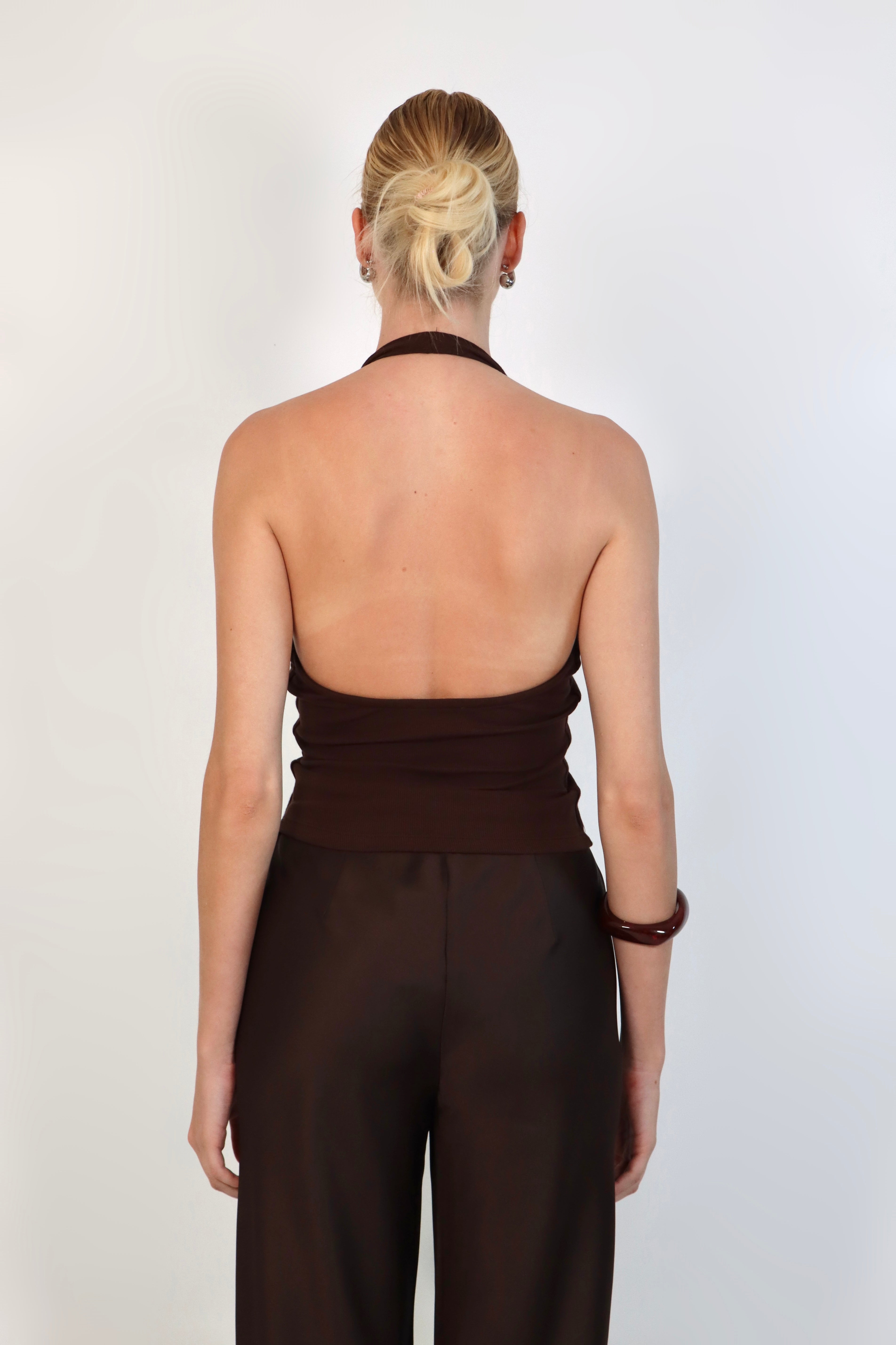 A model wearing a deep Espresso halter top with a ribbed texture and a plunging V-neckline. The fitted top is styled with matching dark brown, high-waisted tailored trousers for a sleek, monochromatic look against a white studio background.