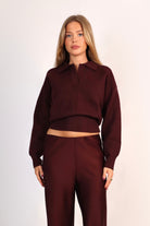 A woman modeling a matching two-piece outfit consisting of a long-sleeve collared crop top and matching wide-leg trousers in a deep burgundy color, set against a plain light background.