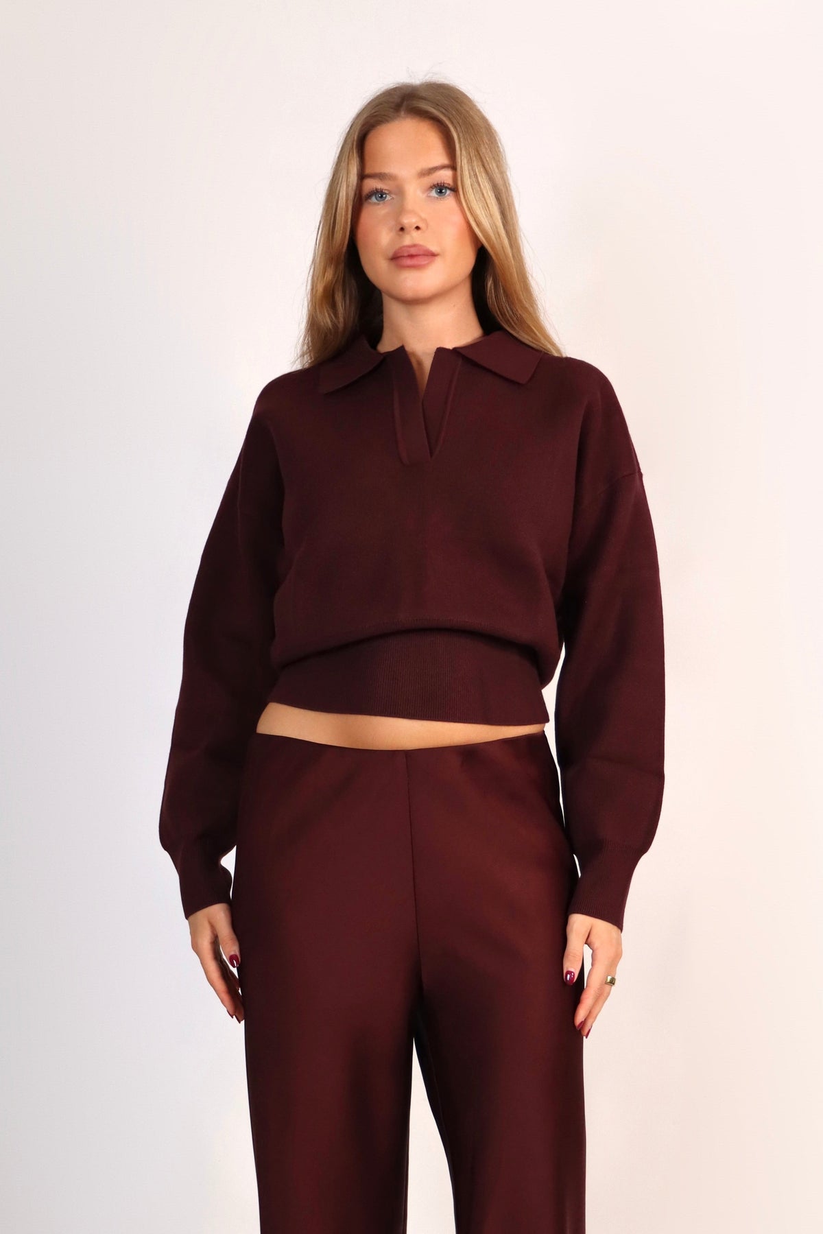 A woman modeling a matching two-piece outfit consisting of a long-sleeve collared crop top and matching wide-leg trousers in a deep burgundy color, set against a plain light background.