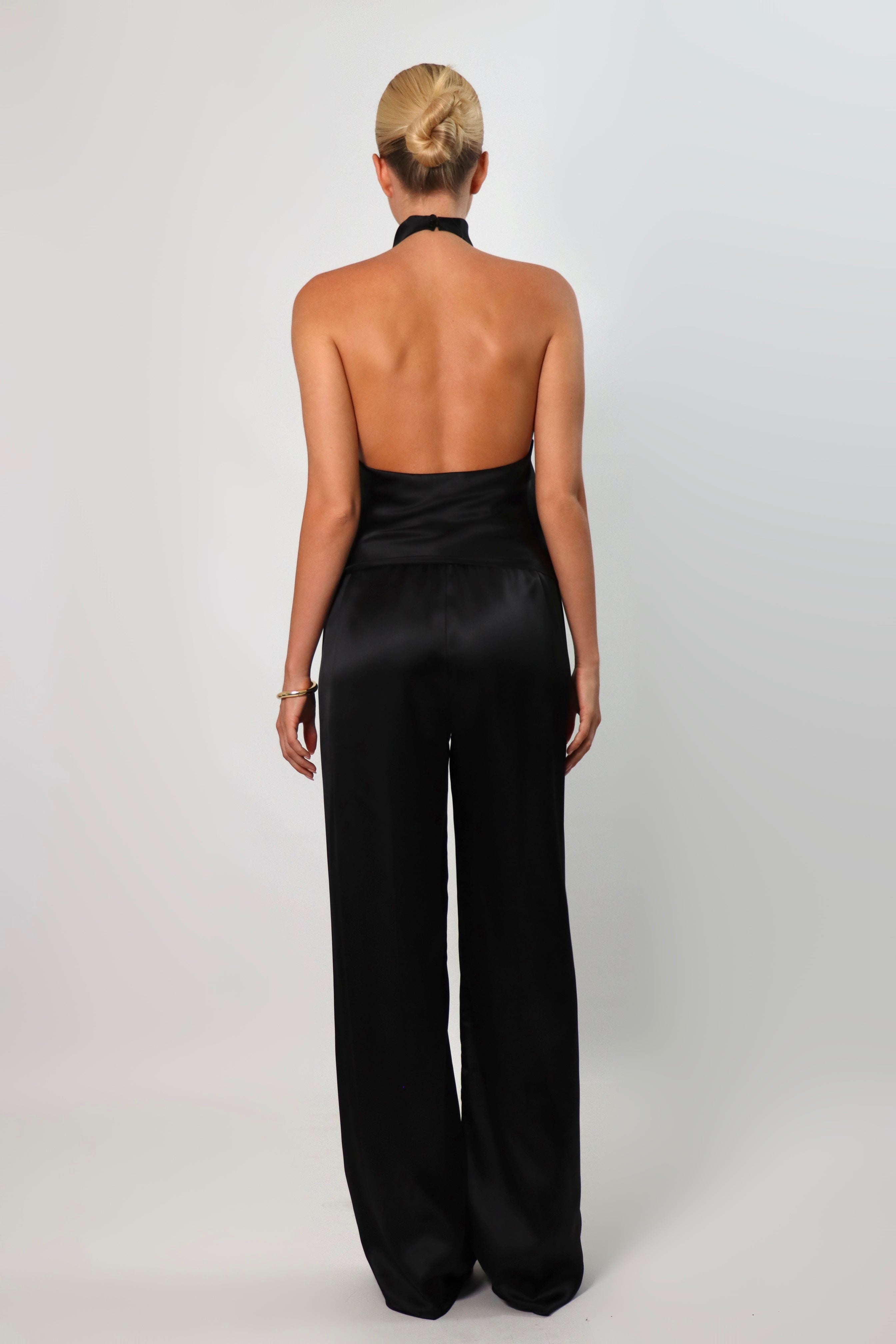 Woman wearing a black satin halter top with a draped neckline, styled with matching satin pants. The top has a smooth, glossy finish and a loose, elegant fit, creating a sophisticated and minimalist look against a light background.