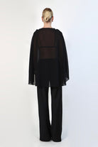  model poses against a neutral background wearing a sheer, black Chiffon Cape Top layered over a matching white bikini top. The cape features an asymmetrical, flowing drape that falls gracefully over high-waisted, cream-colored April Satin Pants and white strappy heels.