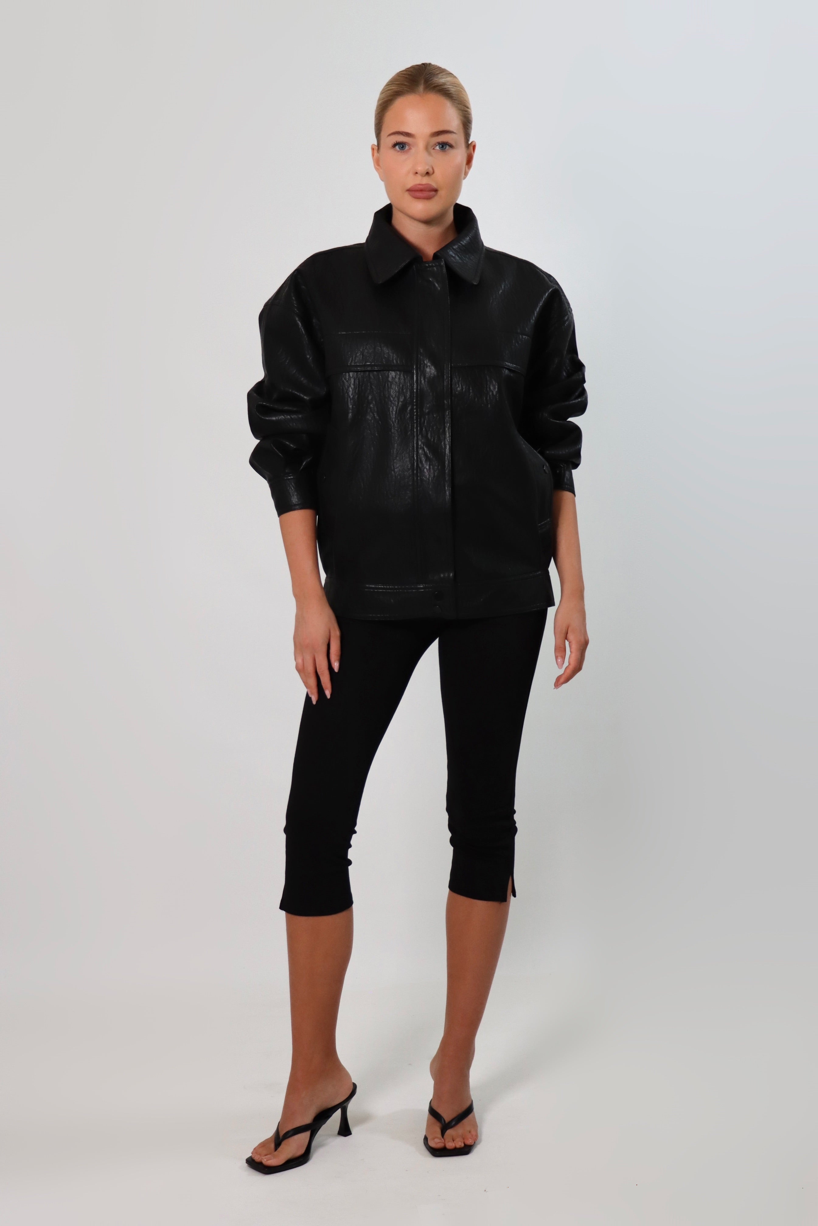 Model wearing a black textured leather oversized jacket with a structured collar and long sleeves, styled with black pants against a neutral background.
