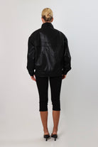 Model wearing a black textured leather oversized jacket with a structured collar and long sleeves, styled with black pants against a neutral background.
