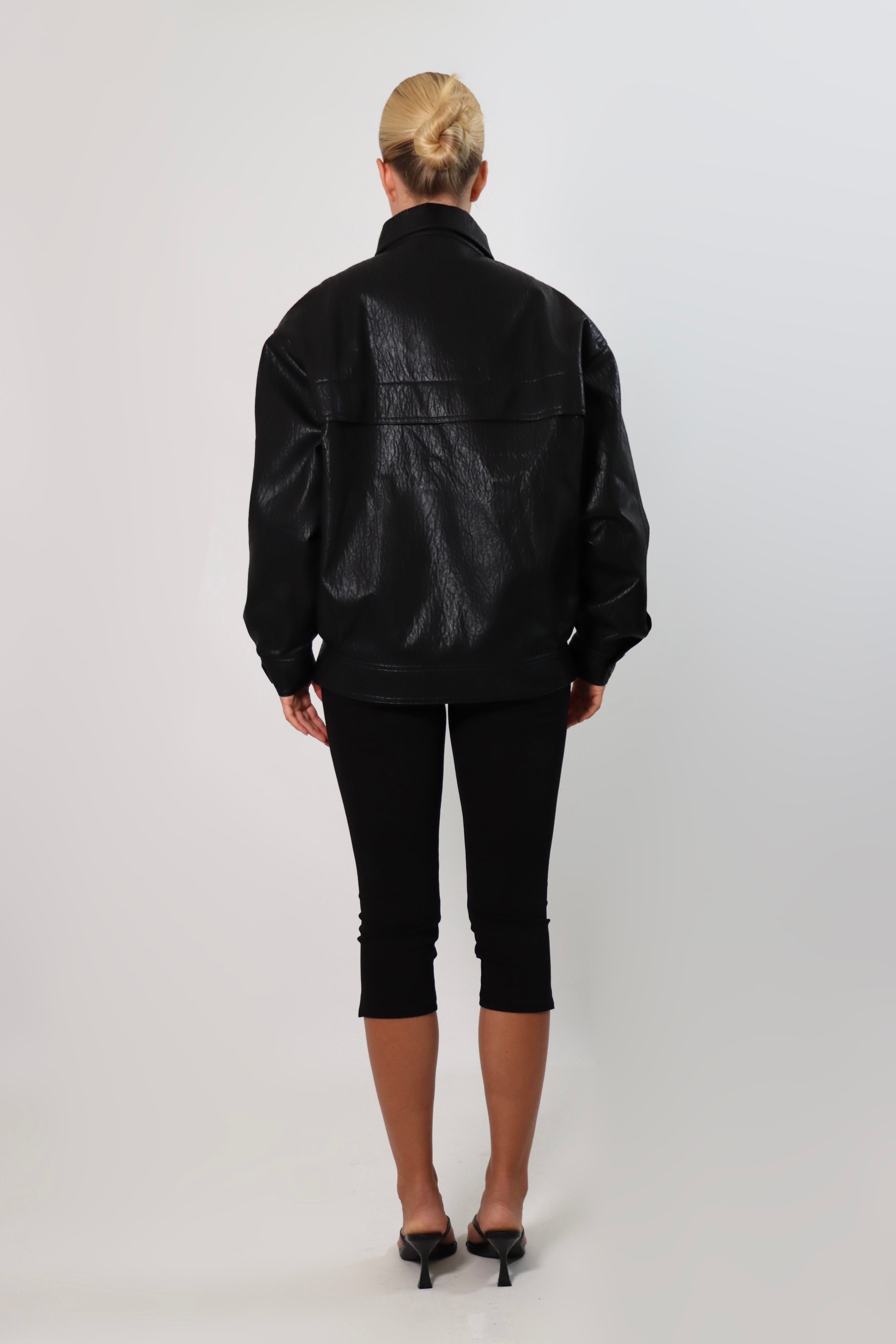 Model wearing a black textured leather oversized jacket with a structured collar and long sleeves, styled with black pants against a neutral background.
