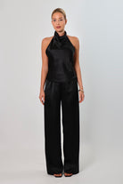 Woman wearing a black satin halter top with a draped neckline, styled with matching satin pants. The top has a smooth, glossy finish and a loose, elegant fit, creating a sophisticated and minimalist look against a light background.