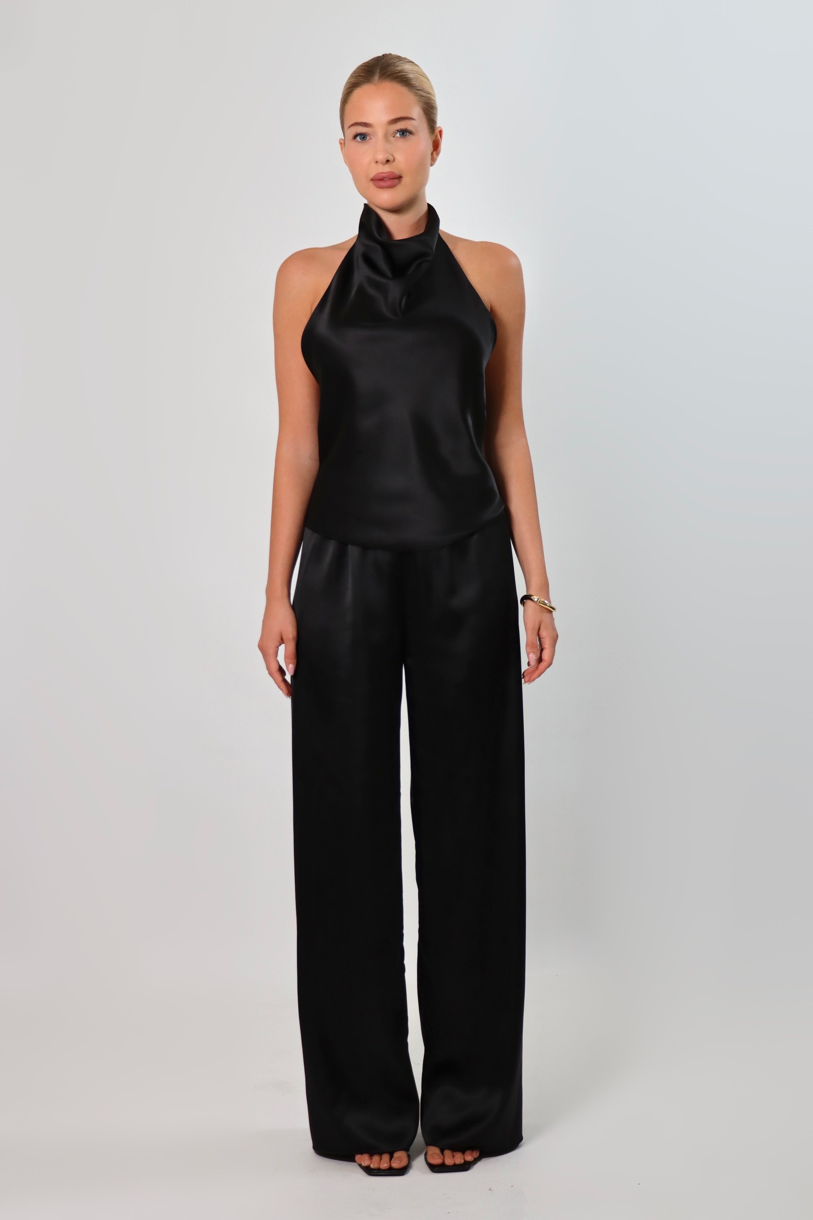 Woman wearing a black satin halter top with a draped neckline, styled with matching satin pants. The top has a smooth, glossy finish and a loose, elegant fit, creating a sophisticated and minimalist look against a light background.