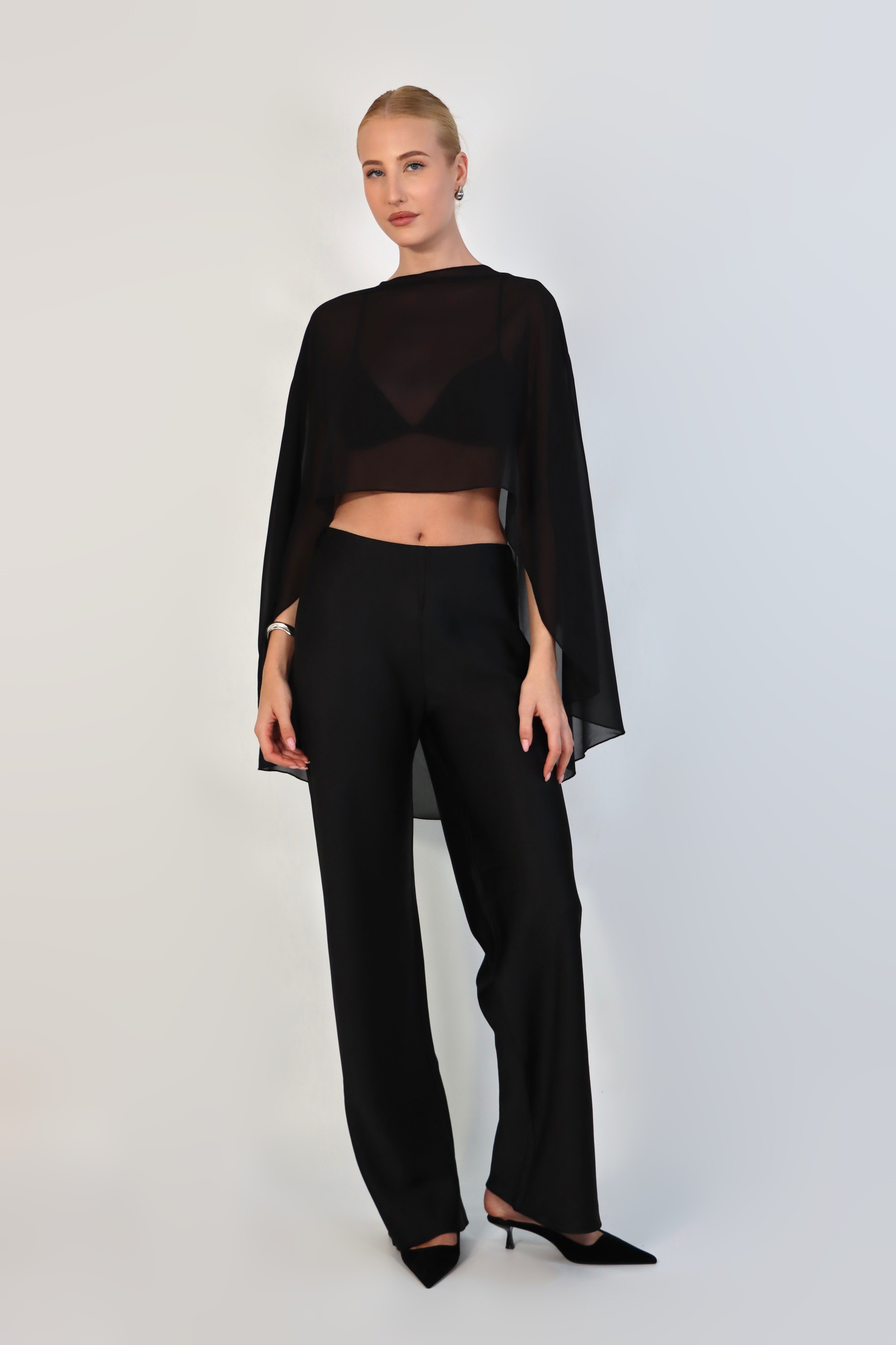  model poses against a neutral background wearing a sheer, black Chiffon Cape Top layered over a matching white bikini top. The cape features an asymmetrical, flowing drape that falls gracefully over high-waisted, cream-colored April Satin Pants and white strappy heels.