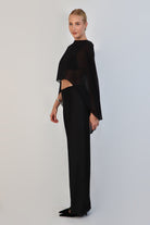  model poses against a neutral background wearing a sheer, black Chiffon Cape Top layered over a matching white bikini top. The cape features an asymmetrical, flowing drape that falls gracefully over high-waisted, cream-colored April Satin Pants and white strappy heels.