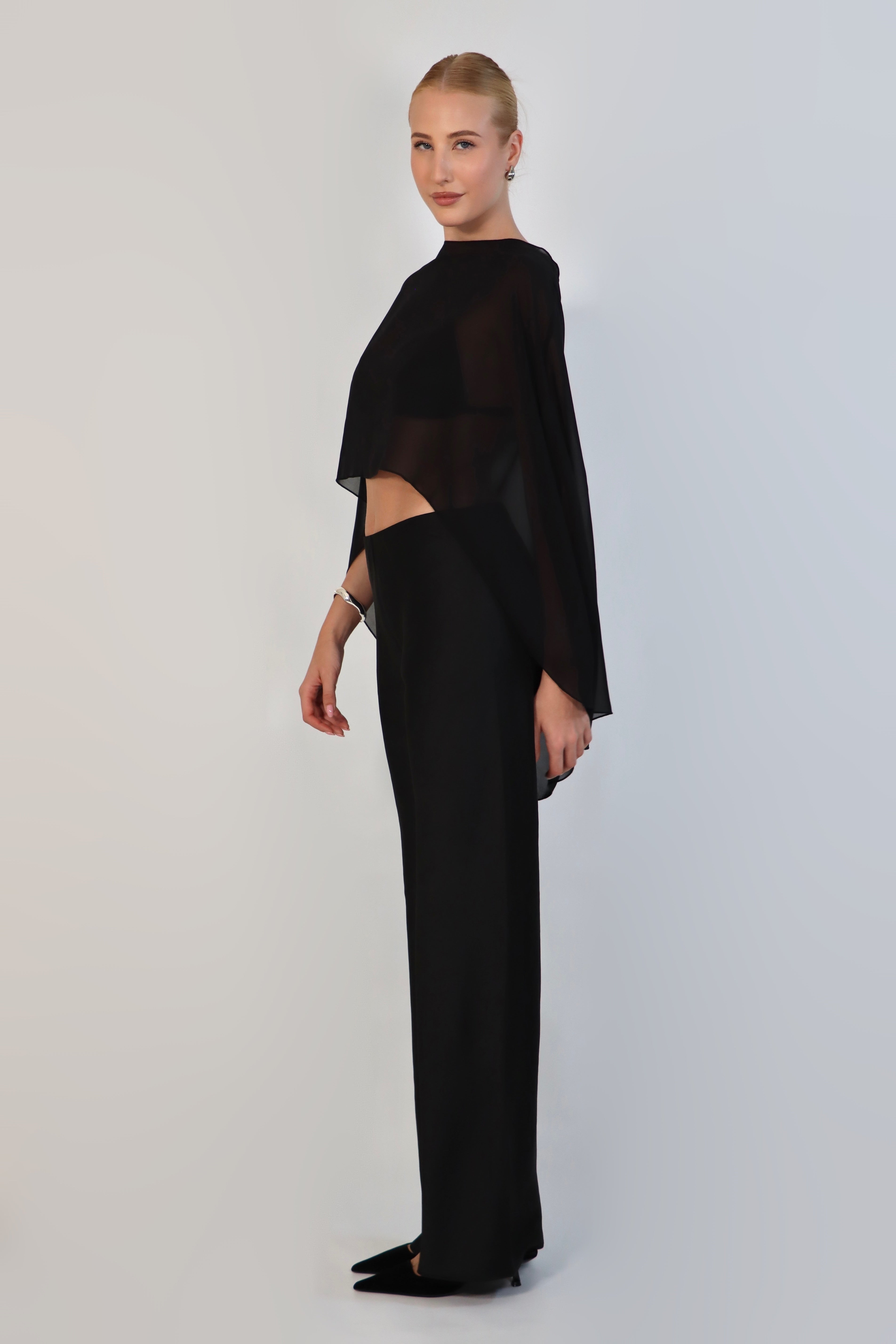  model poses against a neutral background wearing a sheer, black Chiffon Cape Top layered over a matching white bikini top. The cape features an asymmetrical, flowing drape that falls gracefully over high-waisted, cream-colored April Satin Pants and white strappy heels.