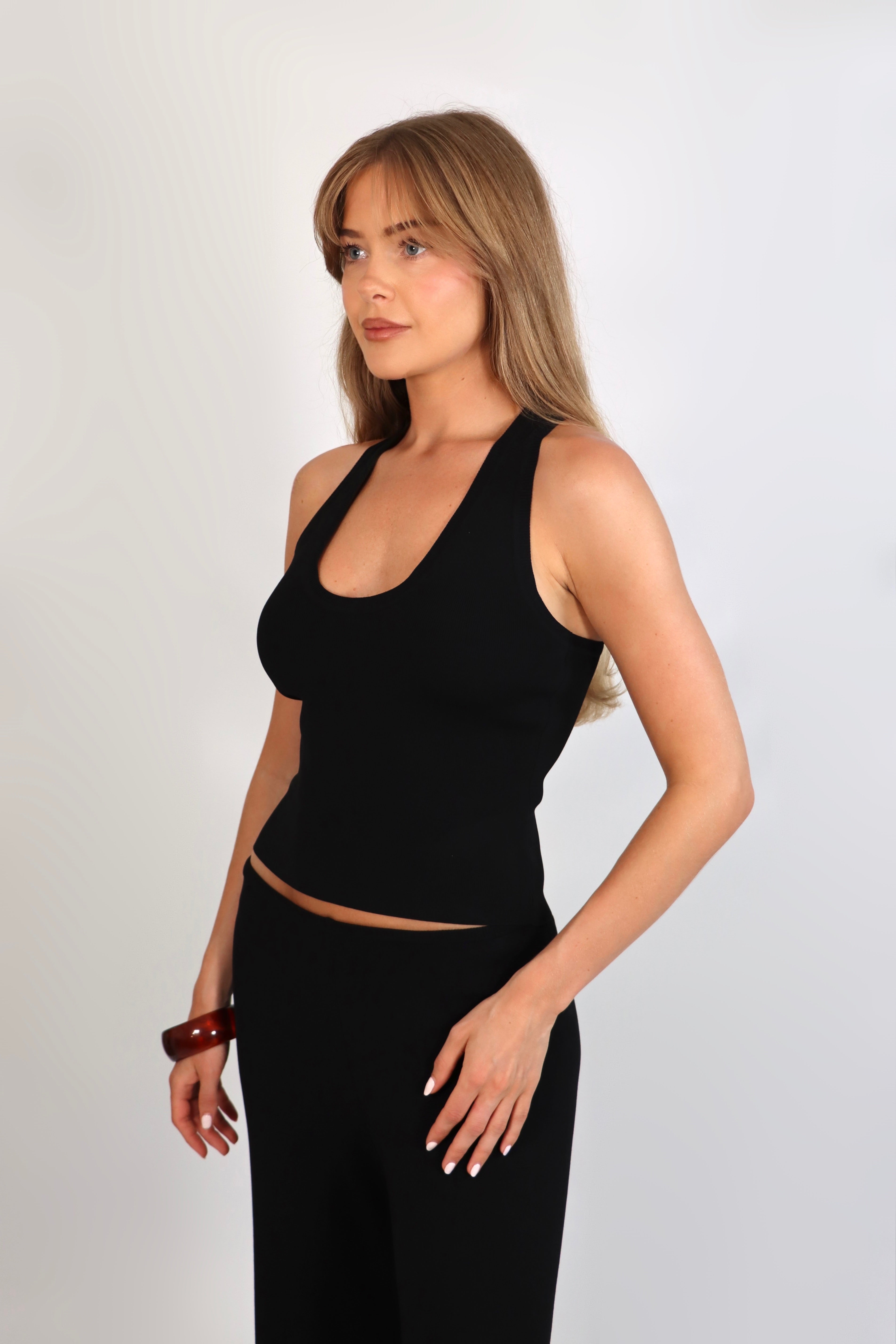 A woman wearing the Freya Tank, featuring a deep scoop neckline and a fitted racerback silhouette. The image highlights the fine rib fabrication and the body-skimming fit of the top in a neutral setting.