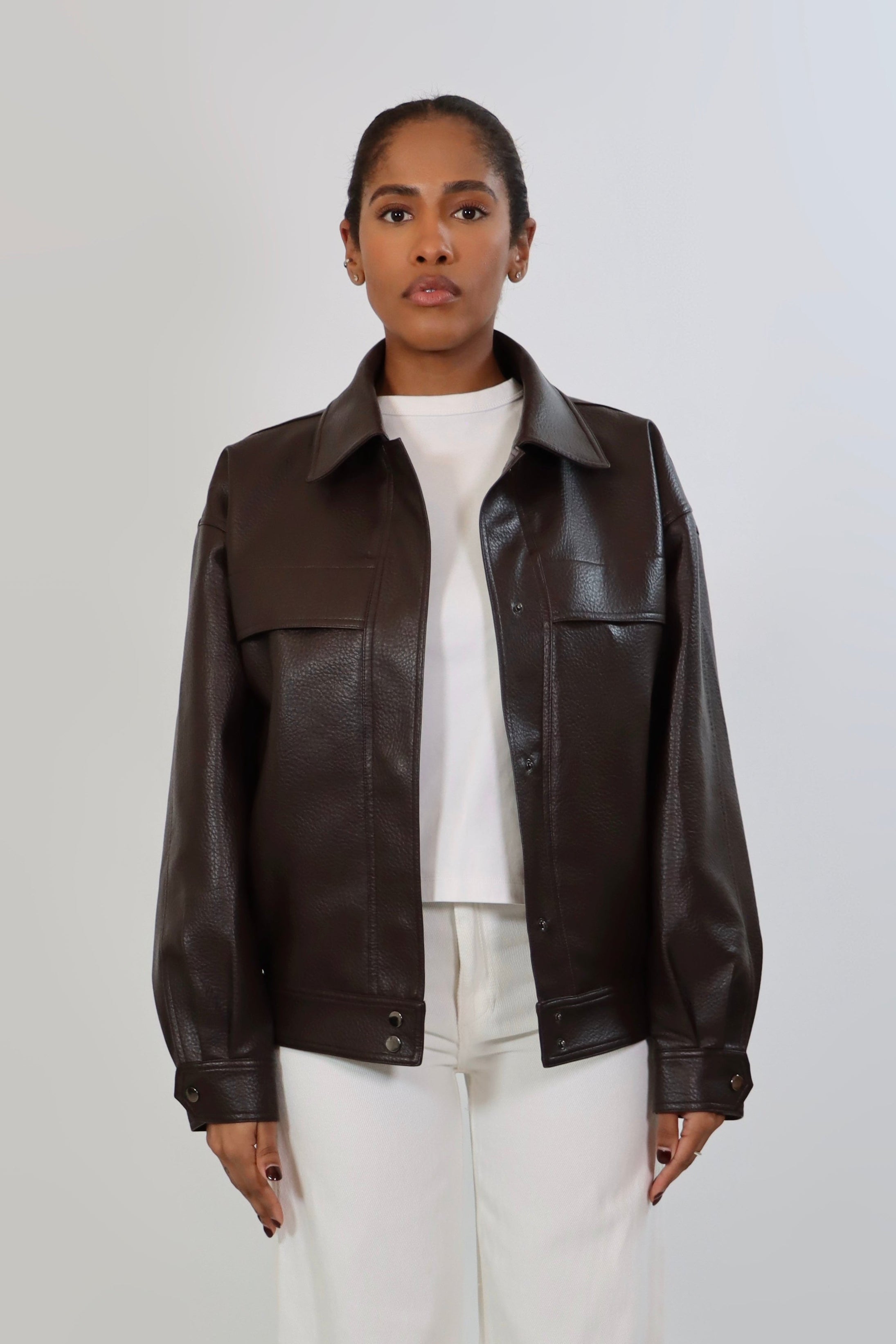 Model wearing an oversized espresso brown faux leather jacket with a boxy fit, spread collar, and front snap closure, styled with white pants against a neutral background.