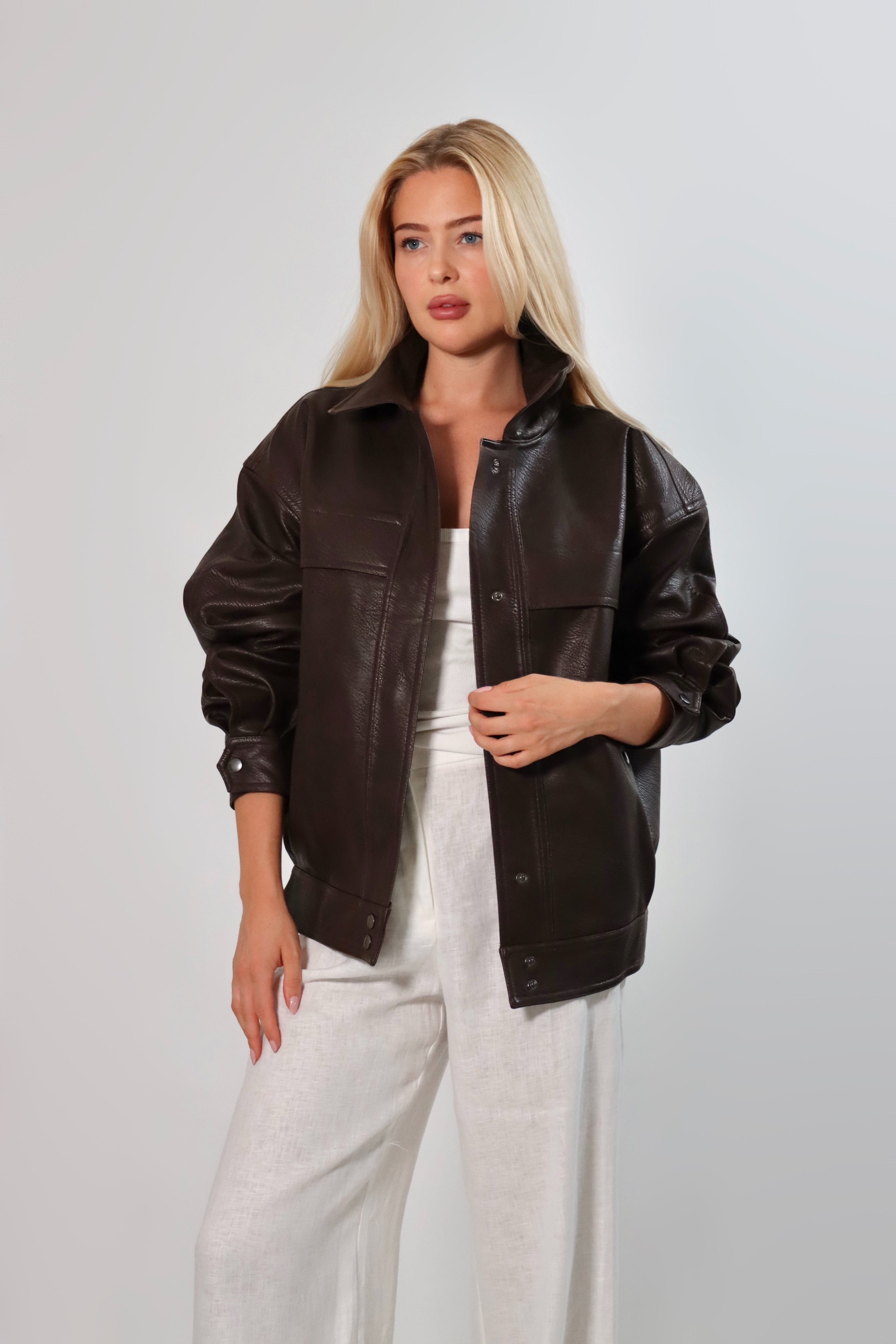 Model wearing an oversized espresso brown faux leather jacket with a boxy fit, spread collar, and front snap closure, styled with white pants against a neutral background.