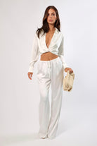Woman in white satin lounge set: long sleeve top and wide leg drawstring pants, with a knit bag.