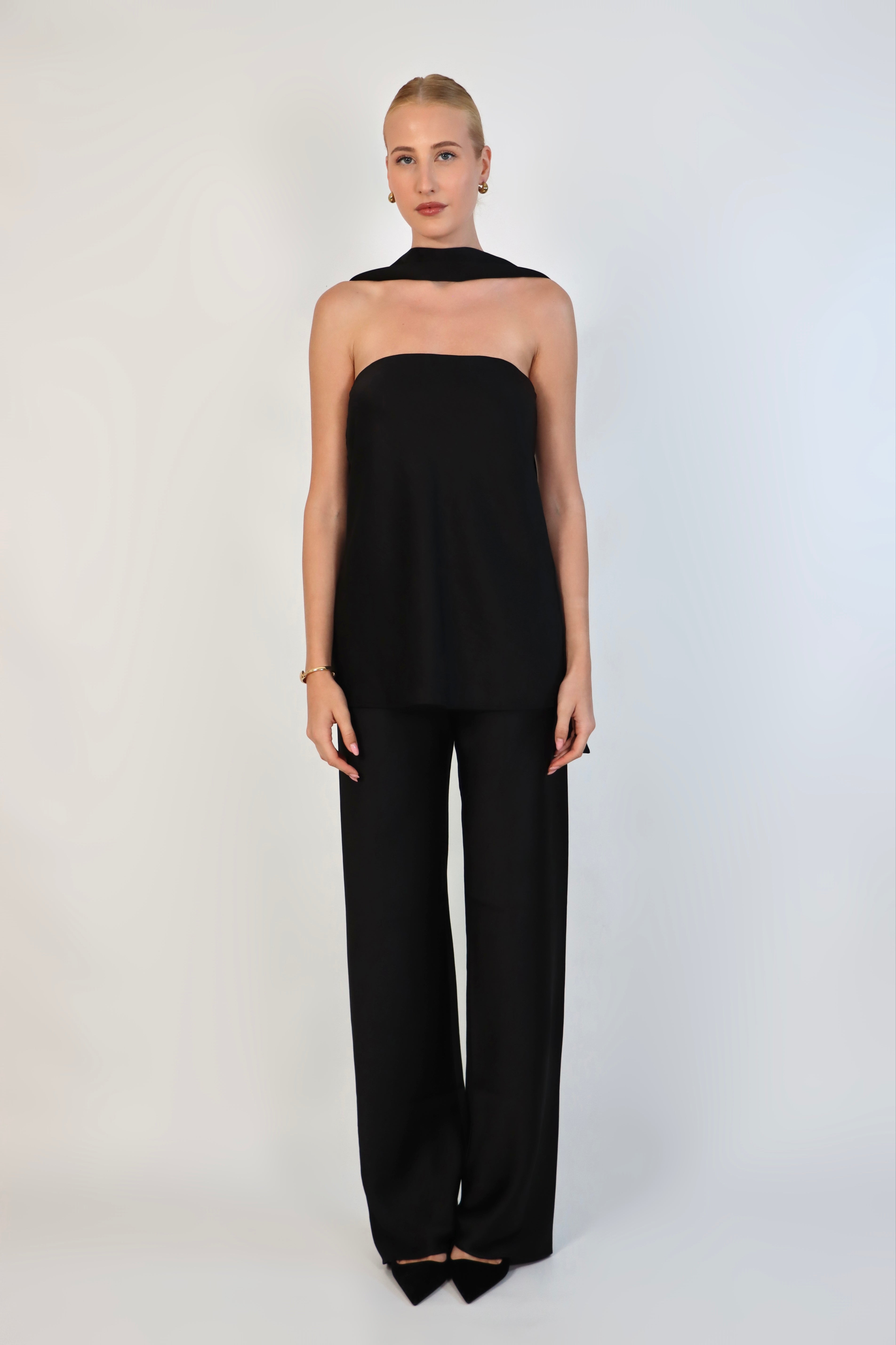 A model wearing a  black, strapless satin top with a matching scarf wrapped elegantly around her neck. The top has a clean, structured bodice and is paired with matching April Satin Pants for a monochromatic, sophisticated look against a plain white background.