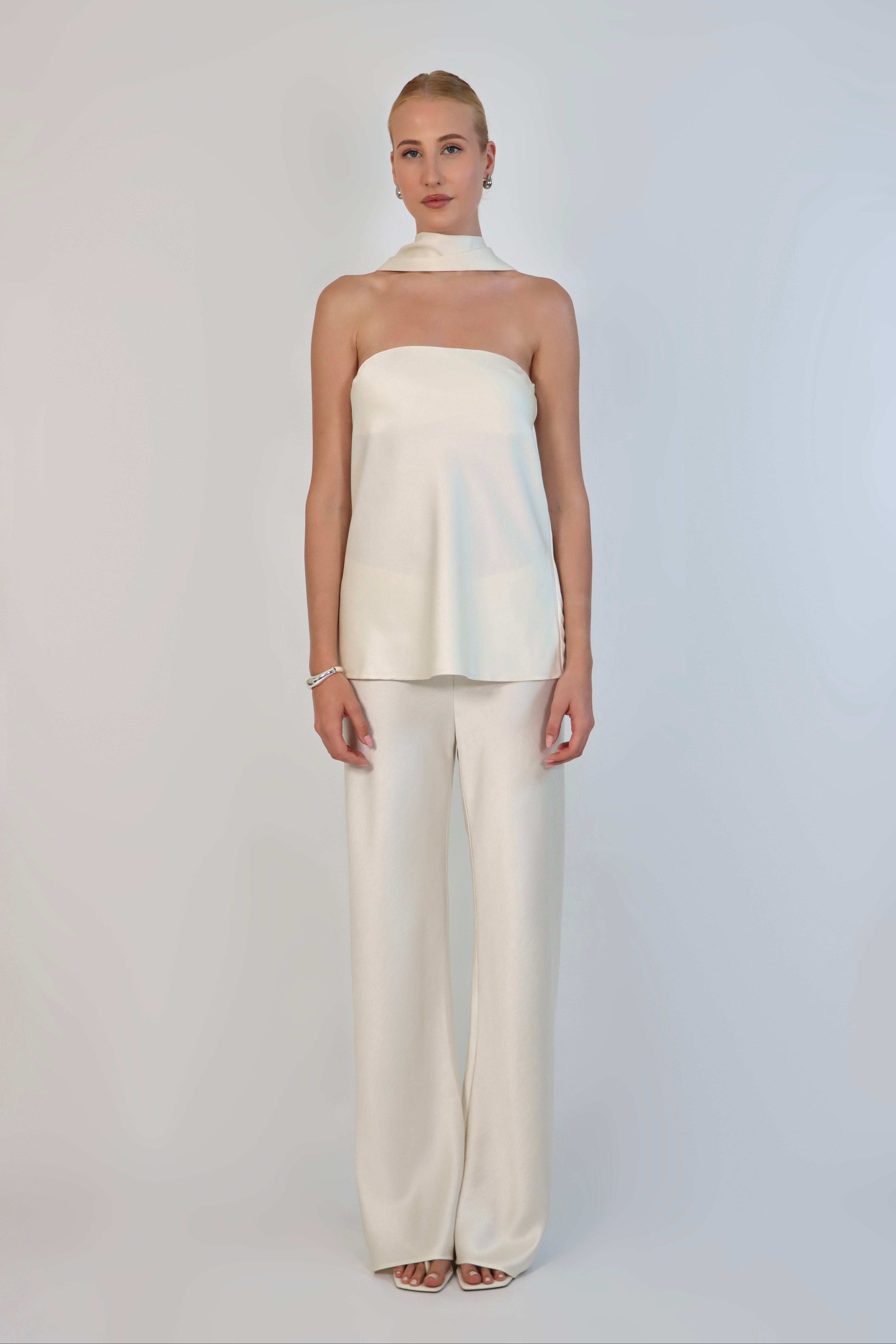 A model wearing a cream-colored, strapless satin top with a matching scarf wrapped elegantly around her neck. The top has a clean, structured bodice and is paired with matching April Satin Pants for a monochromatic, sophisticated look against a plain white background.