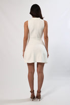 Back view of a woman in a cream sleeveless A-line mini sweater dress with a collar and cinched waist, wearing strappy heels.
