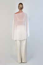 A model poses against a neutral background wearing a sheer, white Chiffon Cape Top layered over a matching white bikini top. The cape features an asymmetrical, flowing drape that falls gracefully over high-waisted, cream-colored April Satin Pants and white strappy heels.