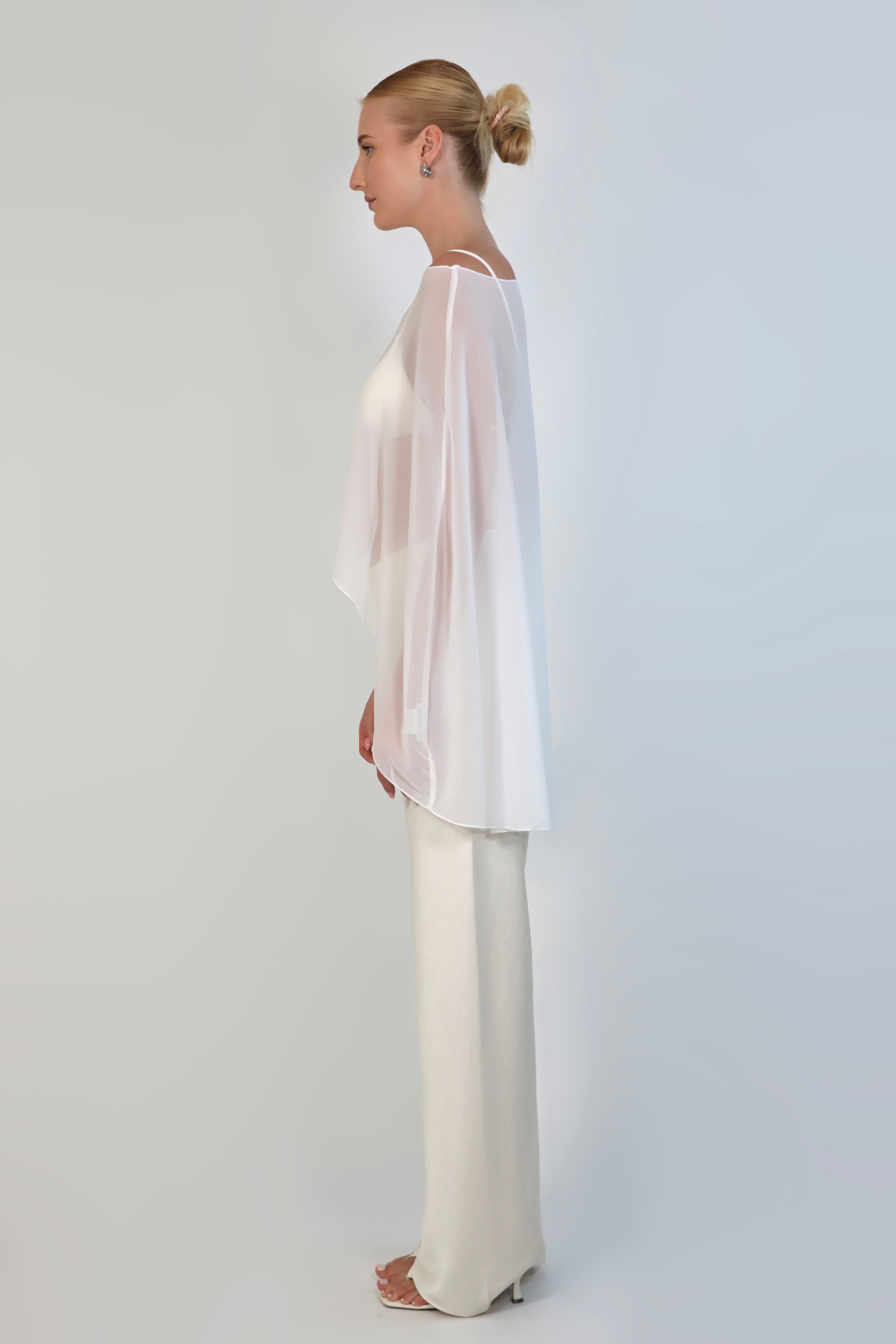 A model poses against a neutral background wearing a sheer, white Chiffon Cape Top layered over a matching white bikini top. The cape features an asymmetrical, flowing drape that falls gracefully over high-waisted, cream-colored April Satin Pants and white strappy heels.